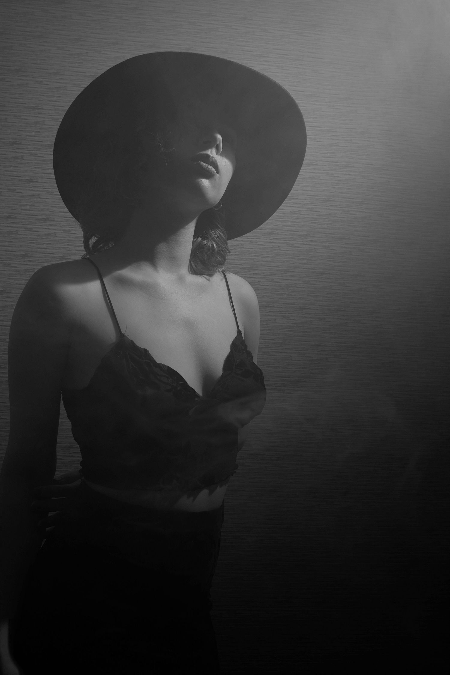 Moody black-and-white portrait of a woman in a wide-brim hat, lit from the side in atmospheric haze.
