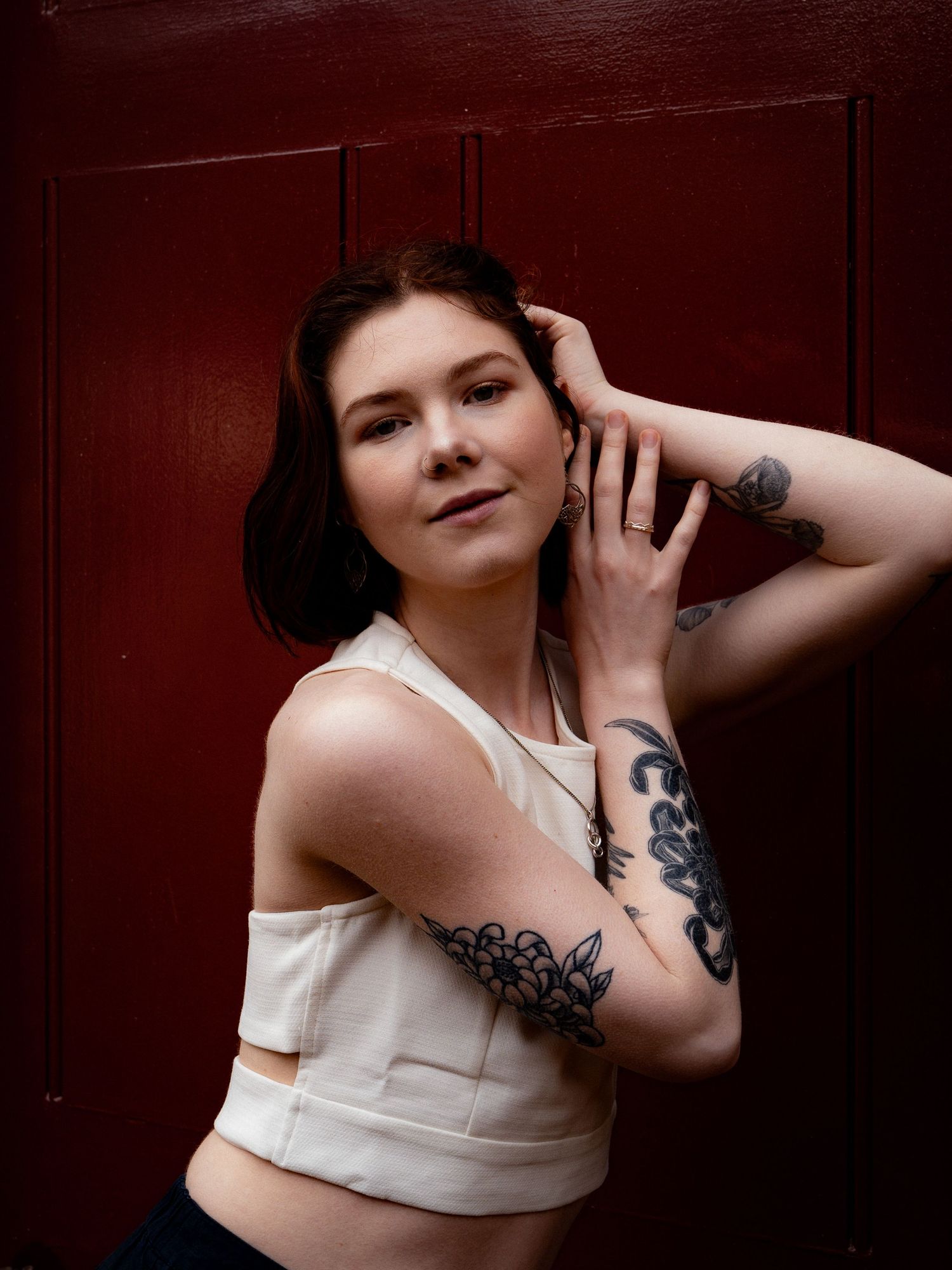 Confident portrait of a woman in a white crop top, leaning against a dark red wall with tattooed arms visible.