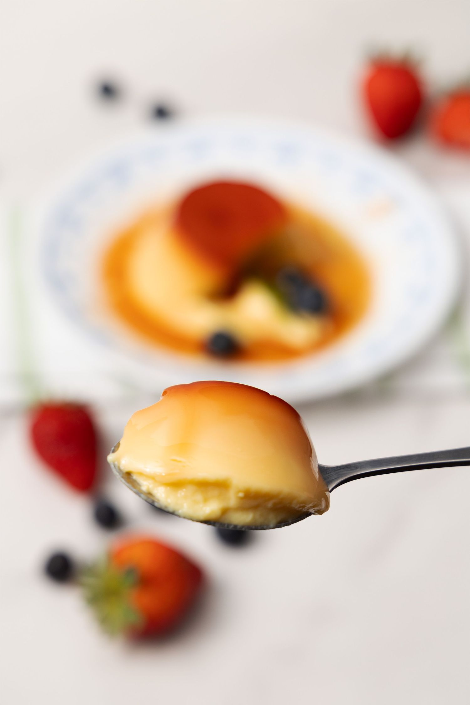 Close-up of a spoonful of custard dessert held in front of a plated flan with strawberries and blueberries.