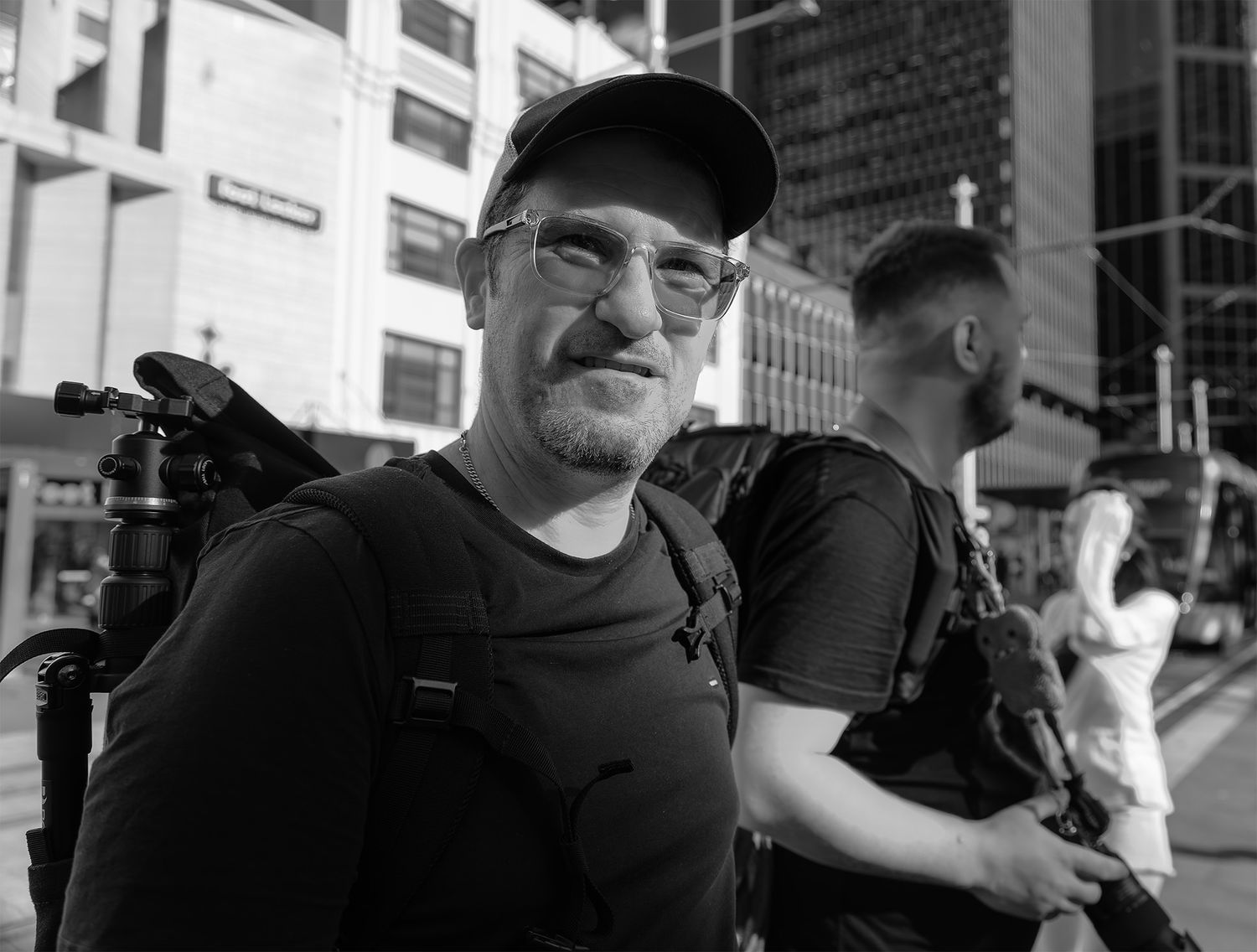 Black-and-white street portrait of a man in cap and glasses with camera backpack, standing in a busy city intersection.