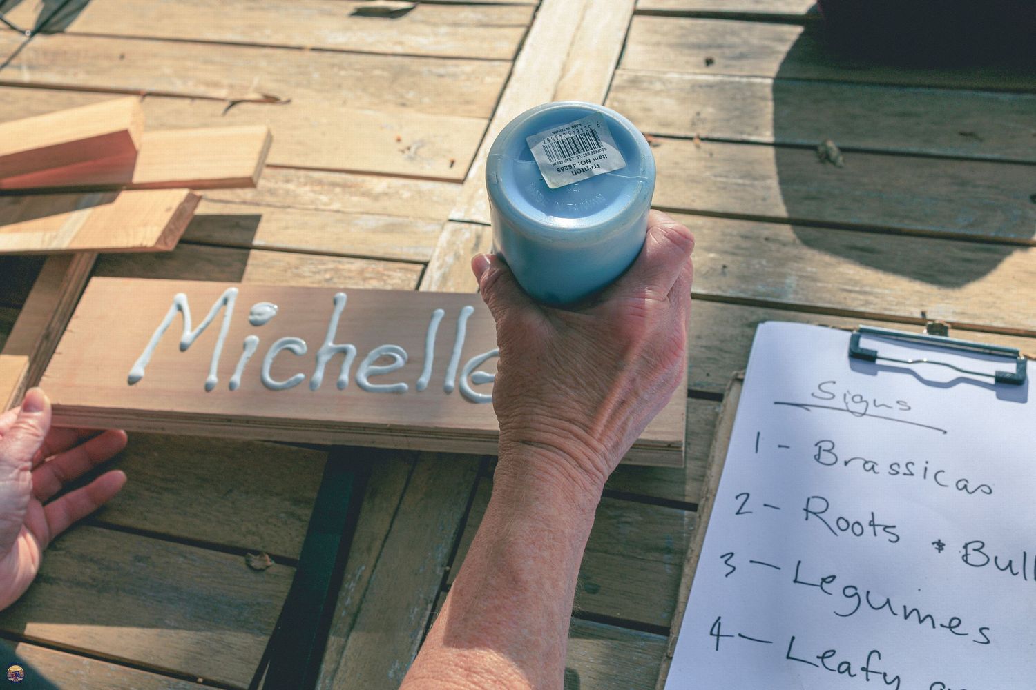 Person writing ‘Michelle’ with white glue on a wooden garden sign, with a planting plan on a clipboard nearby.