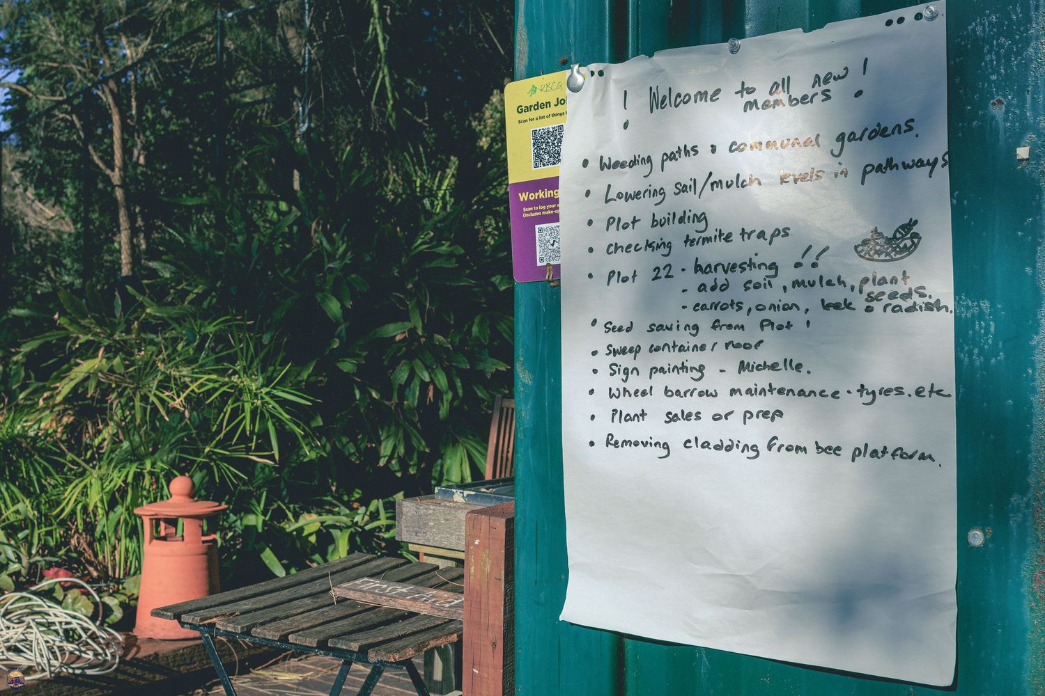 Handwritten meeting notes on butcher’s paper taped to a green shed wall in a community garden, with plants and outdoor furniture behind.