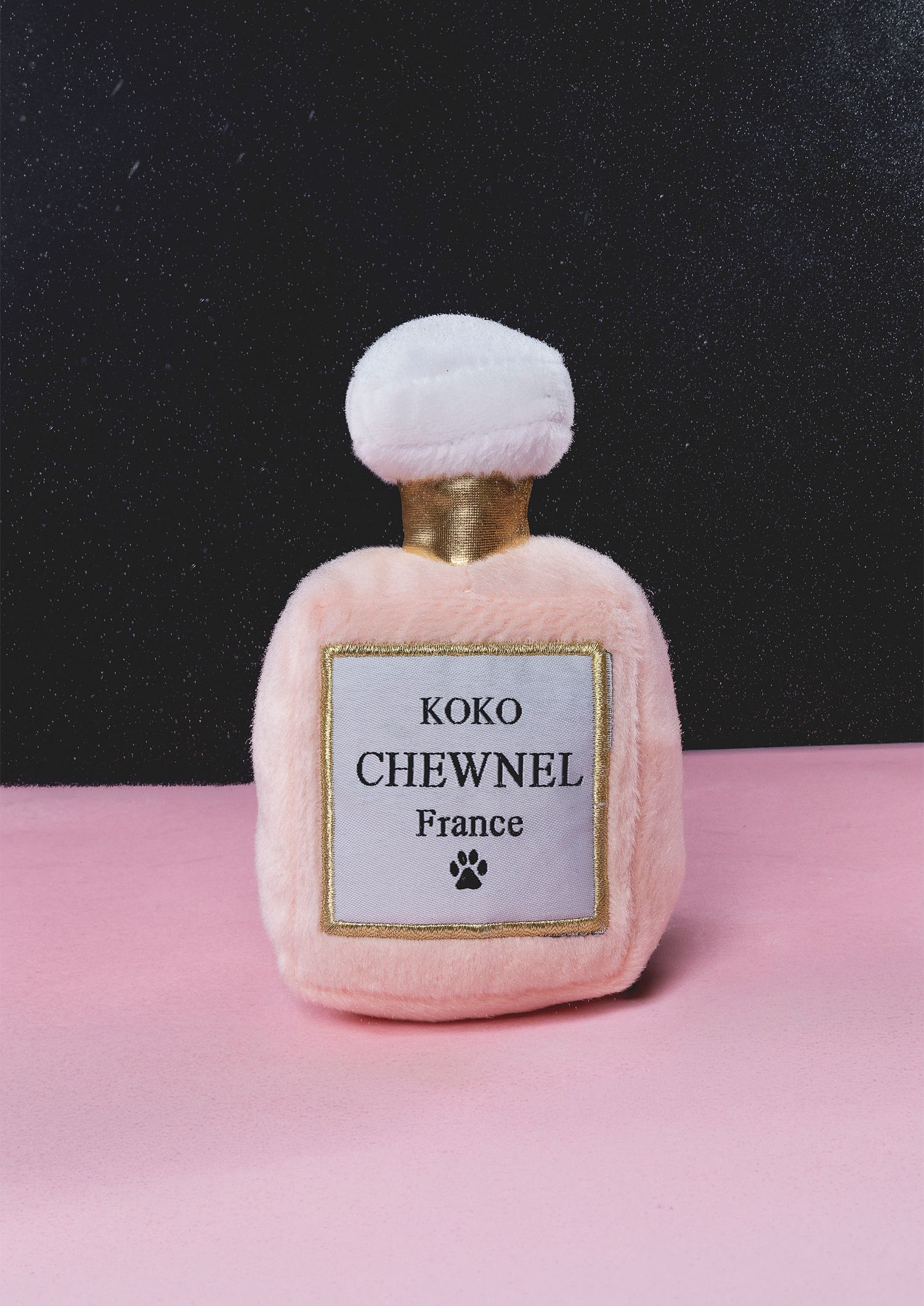 Studio product shot of a plush perfume bottle dog toy on a pink surface against a black background.