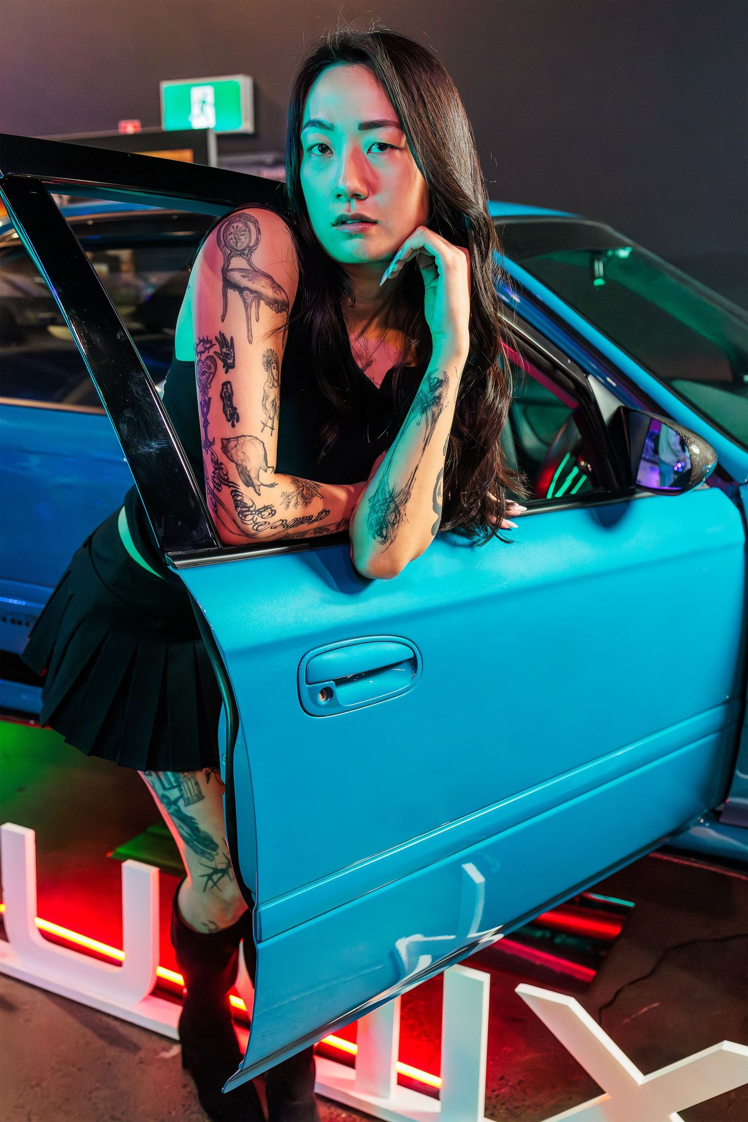 Neon-lit portrait of a tattooed woman leaning on the open door of a blue car at an indoor automotive event.