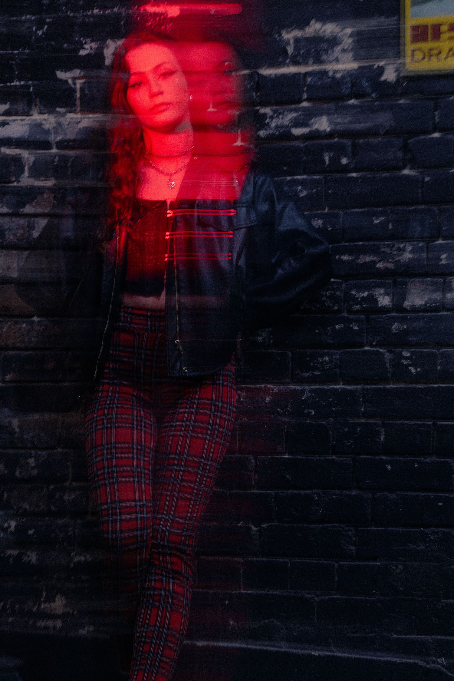 Long-exposure portrait of a woman leaning against a brick wall, with red motion blur creating a ghosted double image.
