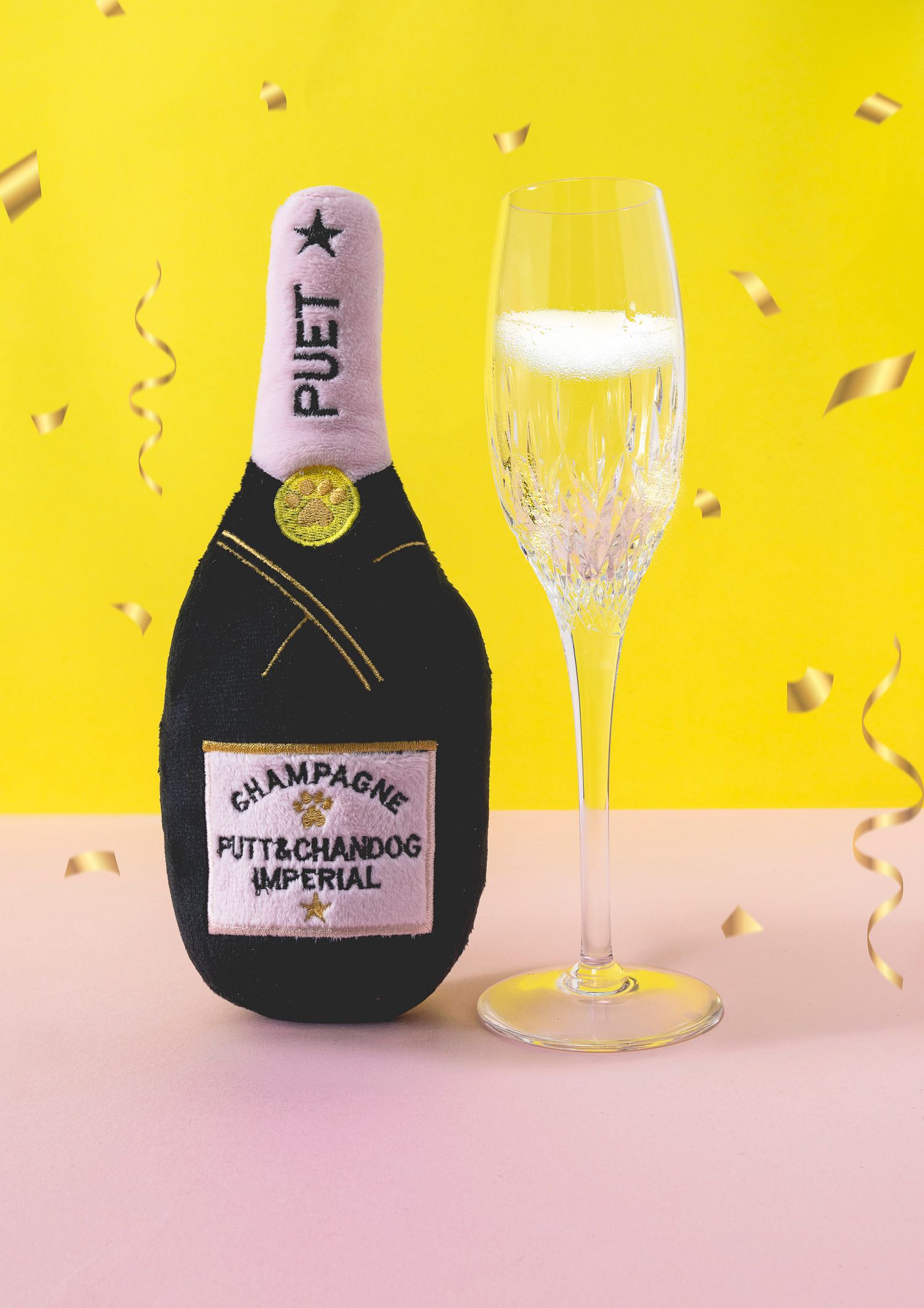 Playful product shot of a plush champagne bottle dog toy and real champagne flute against a bright yellow background.
