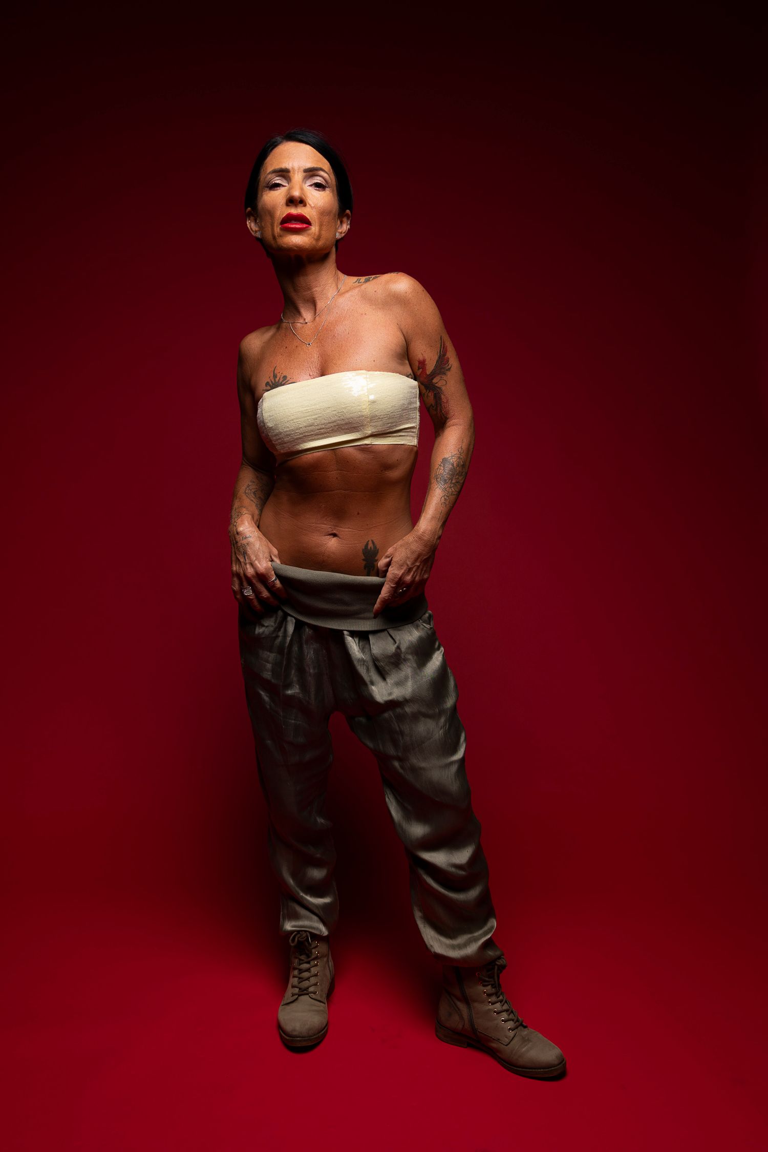Full-body studio portrait of a tattooed woman in a white bandeau top and camo pants, standing confidently against a deep red background.
