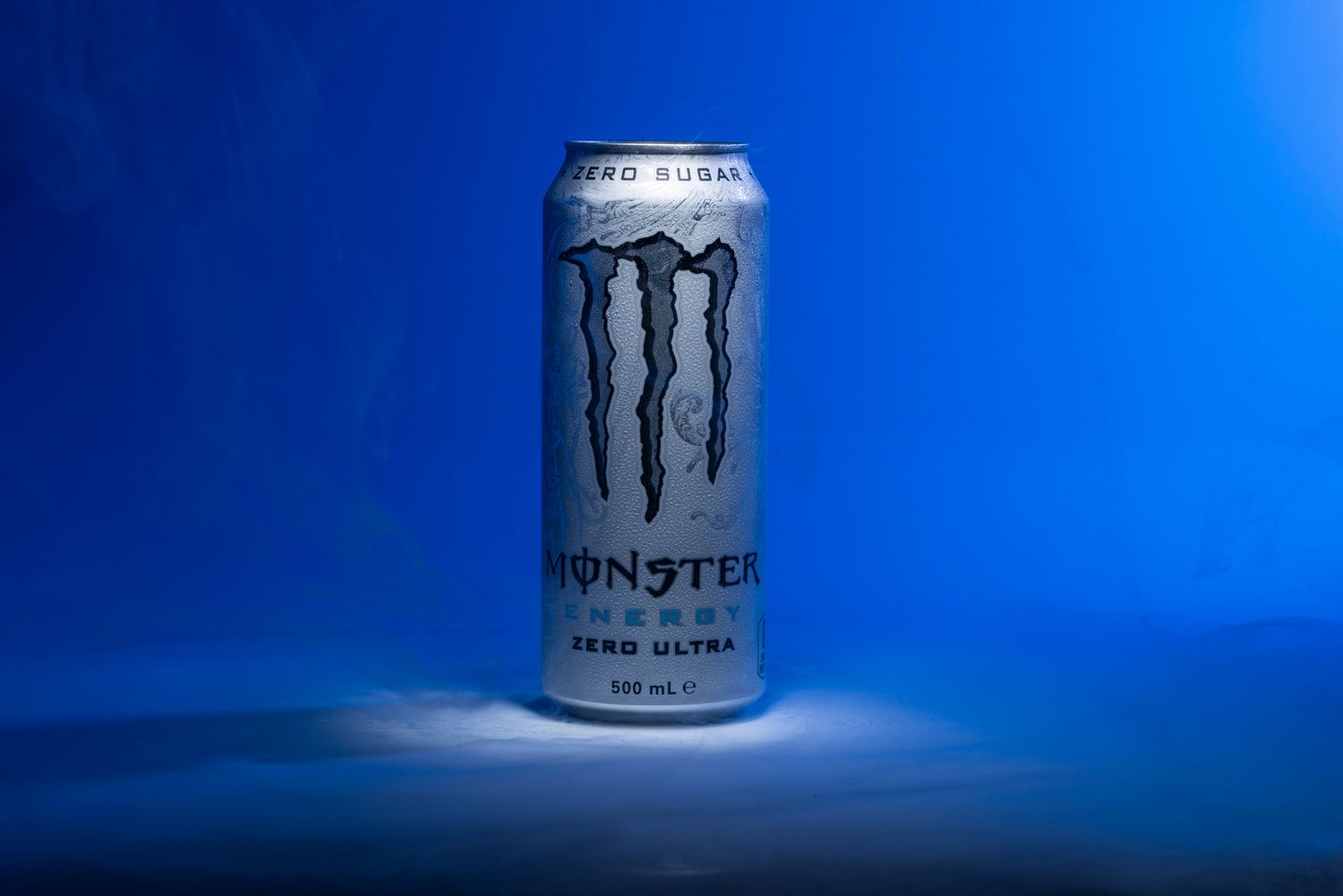Studio product shot of a single Monster Energy can standing on a reflective surface against a vivid blue background.