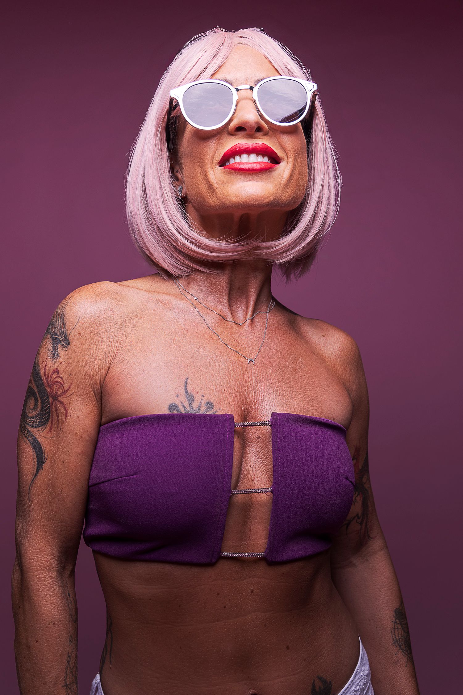 Bold studio portrait of a woman with a pink bob, round sunglasses and purple bandeau top against a magenta background.