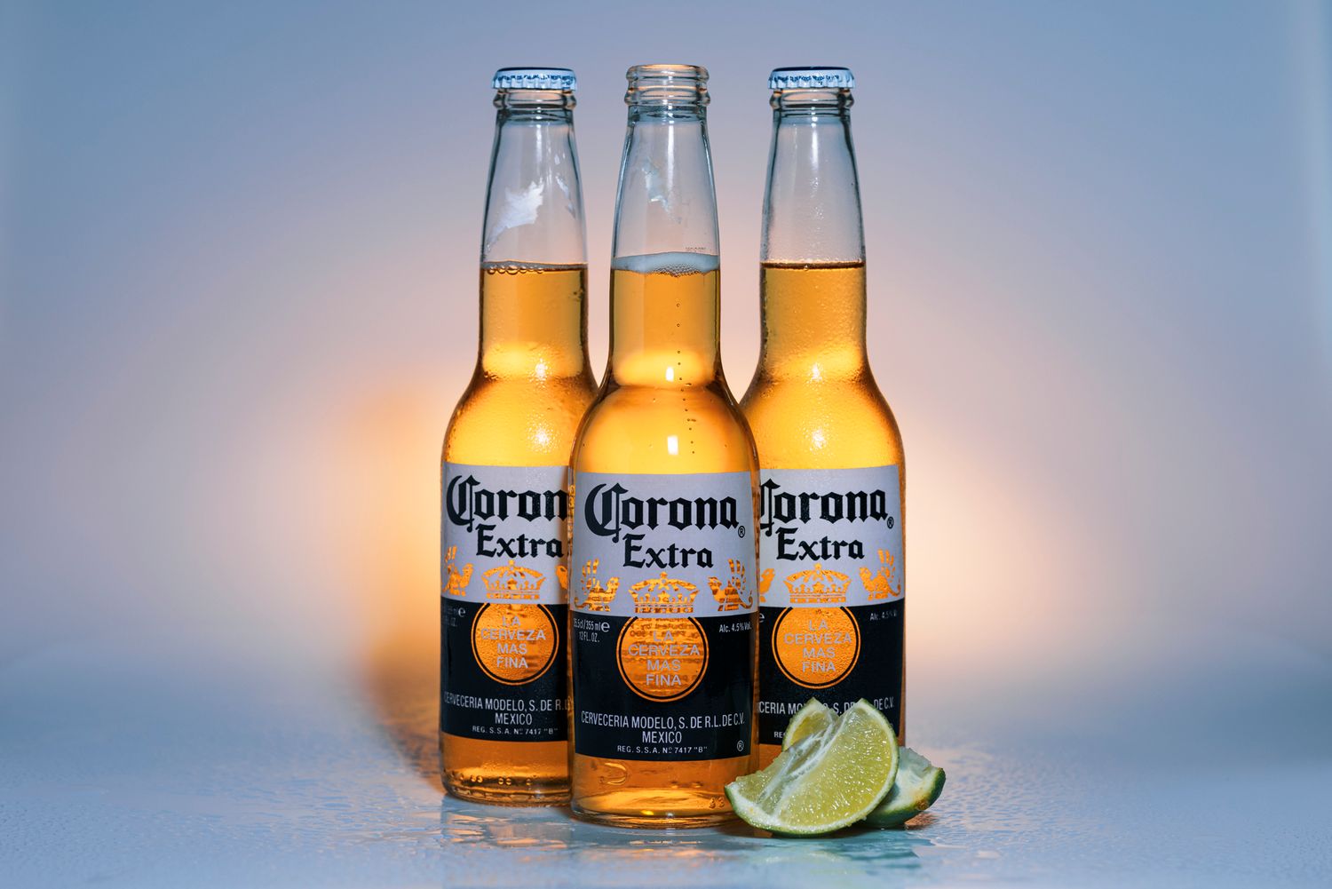 Studio product shot of three Corona beer bottles with lime wedges in front on a cool blue background.