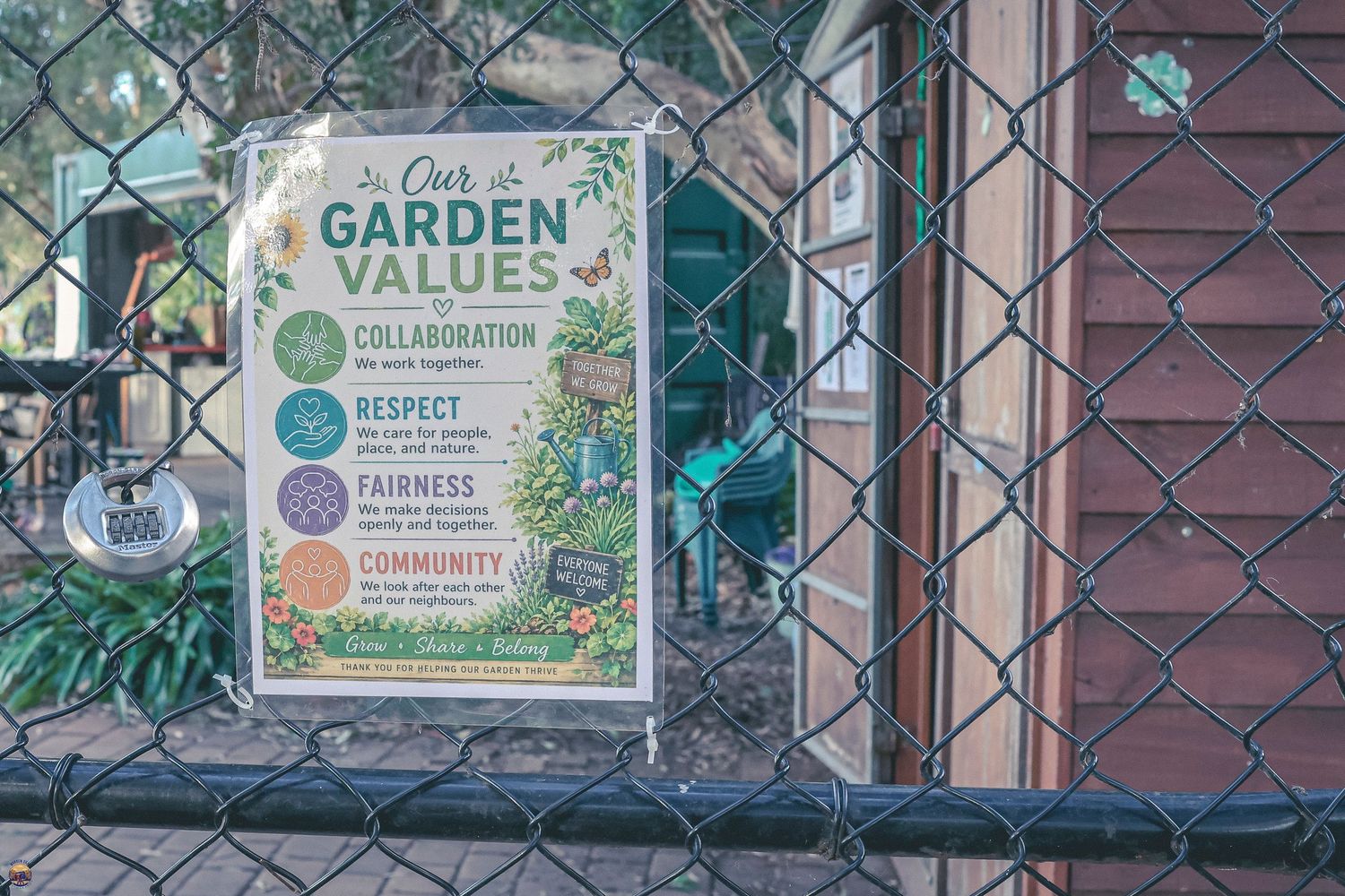 Colourful garden values sign on a chain-link fence at the entrance to a community garden, with sheds and foliage in the background.