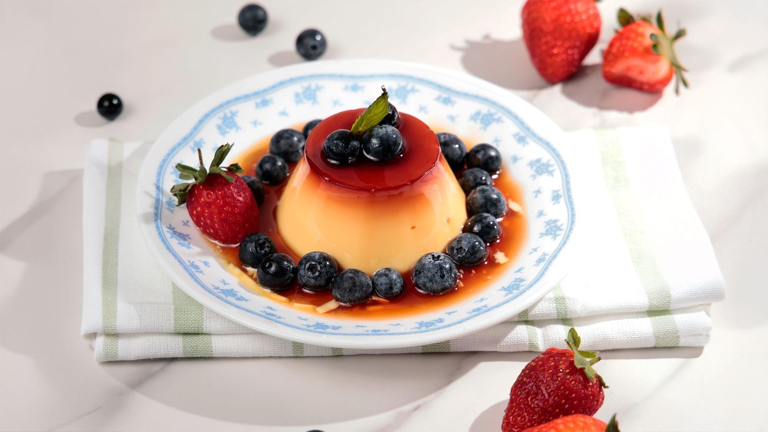 Studio food photograph of a flan-style dessert topped with strawberry and blueberries on a patterned plate.