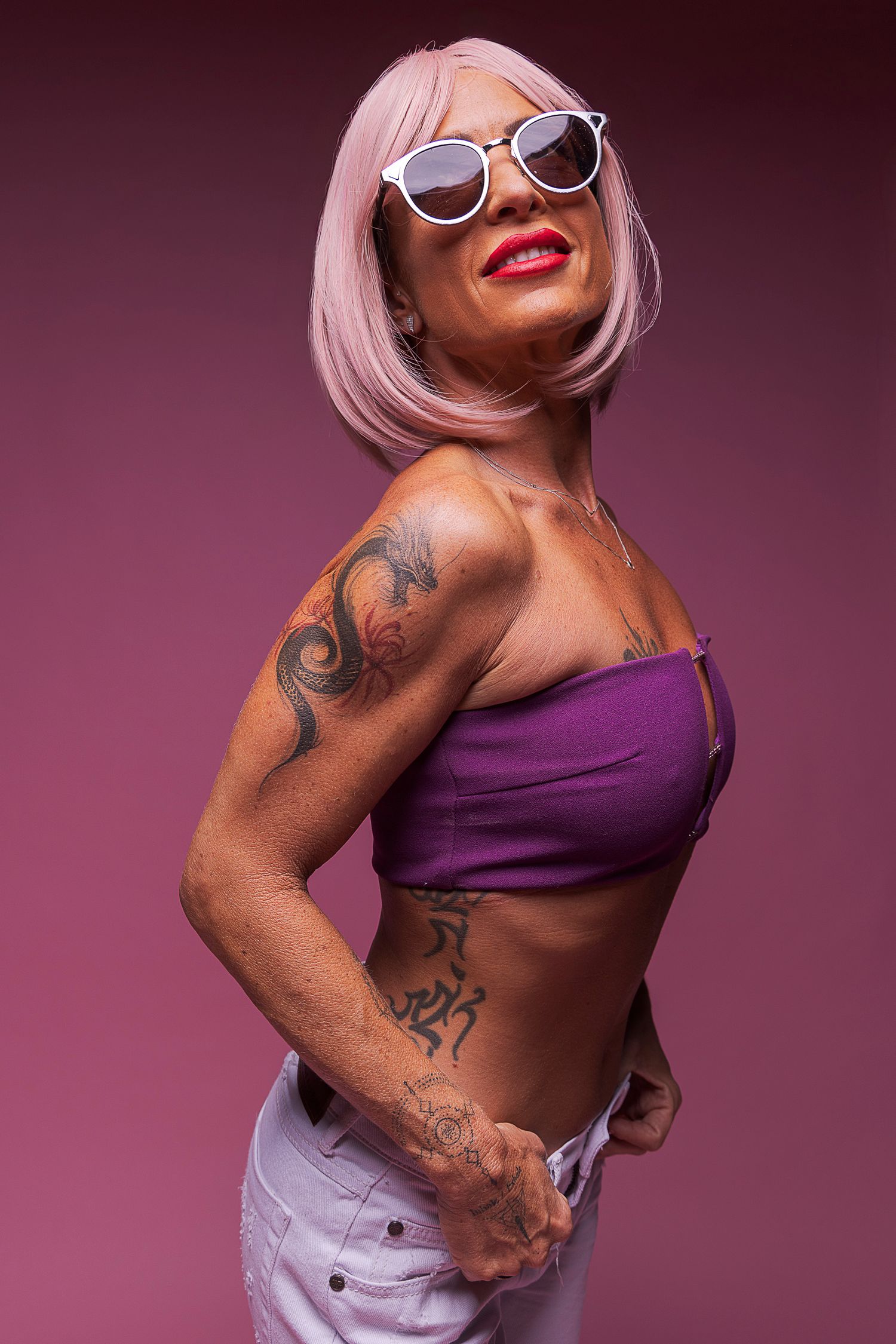 Studio portrait of a woman with pink bob haircut and sunglasses, smiling confidently in a purple bandeau top and white pants against a magen