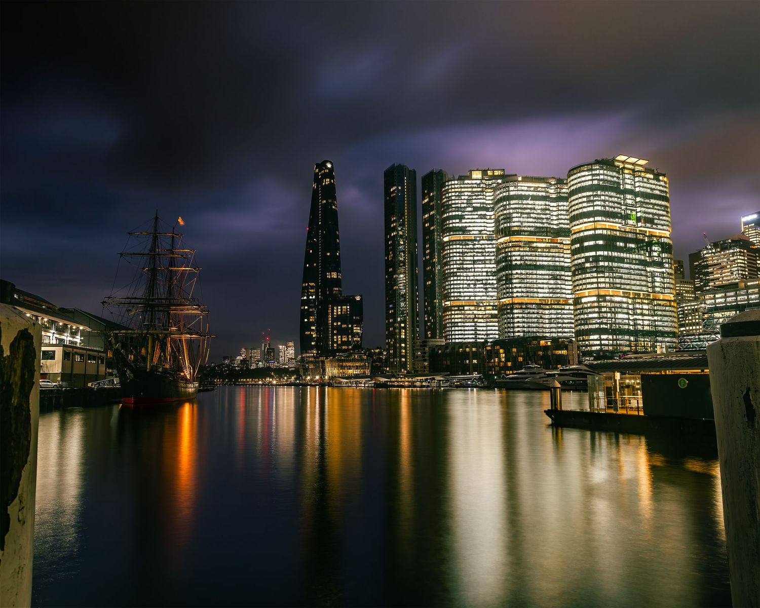 Night-time cityscape of illuminated waterfront office towers reflected in calm harbour water under a dramatic cloudy sky.
