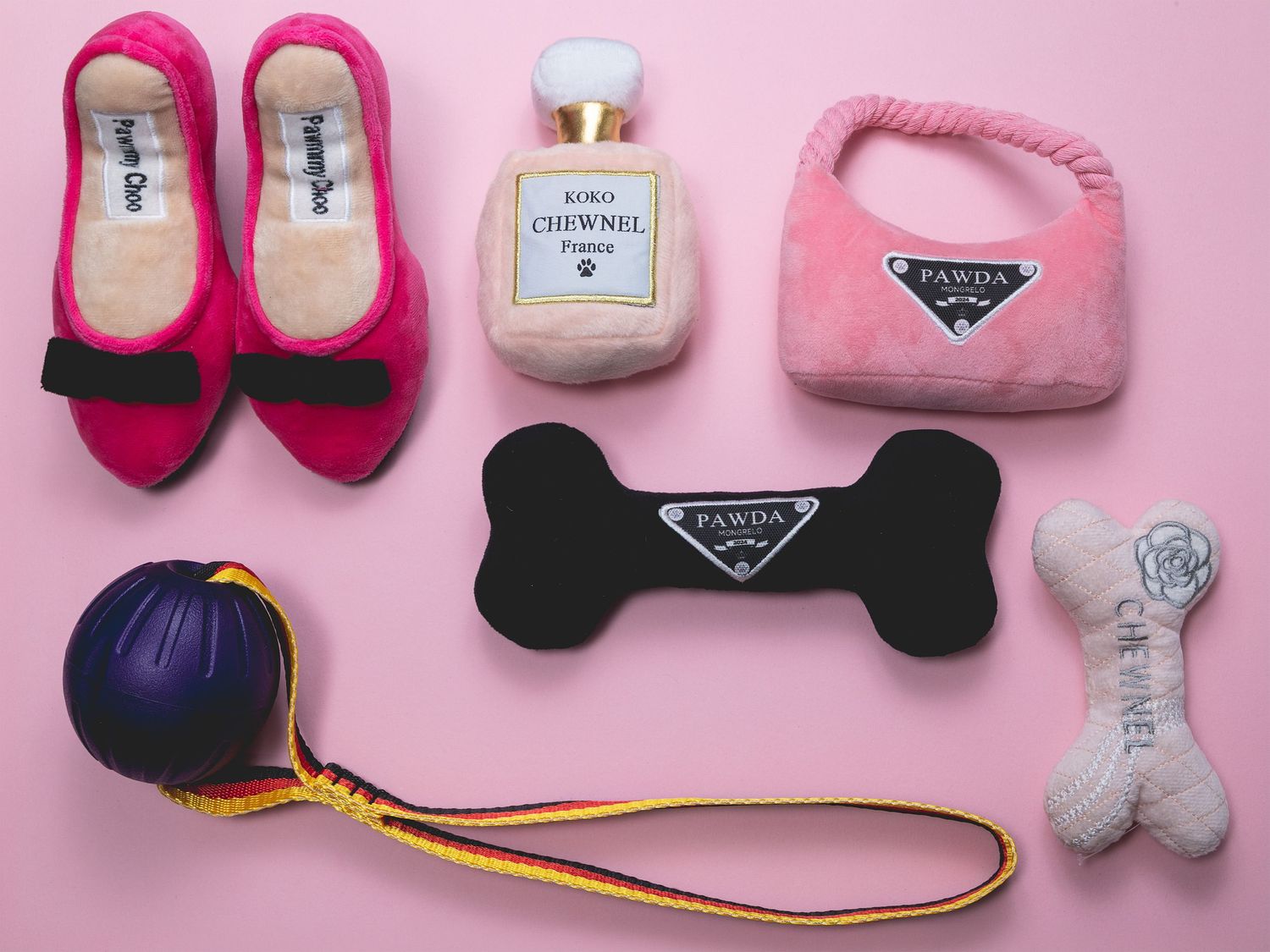 Flat-lay of luxury-inspired dog toys arranged on a pink background, including shoes, perfume bottle, bag, bones and ball.