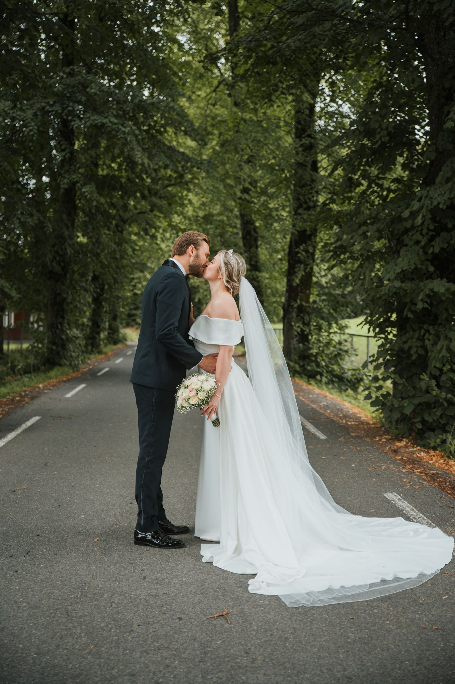 Bride and groom sharing a kiss on scenic tree-lined road, bride in off-shoulder gown with cathedral veil.