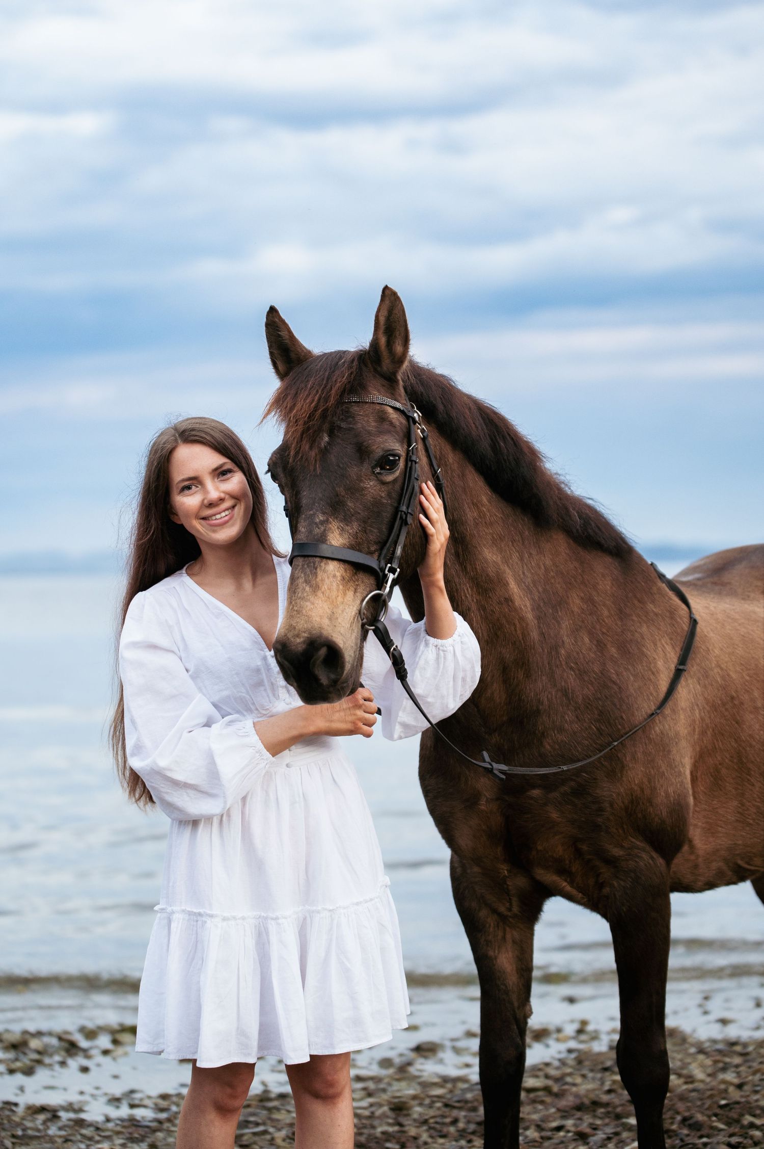Smiling woman in white dress stands beside a brown horse on a sandy beach.