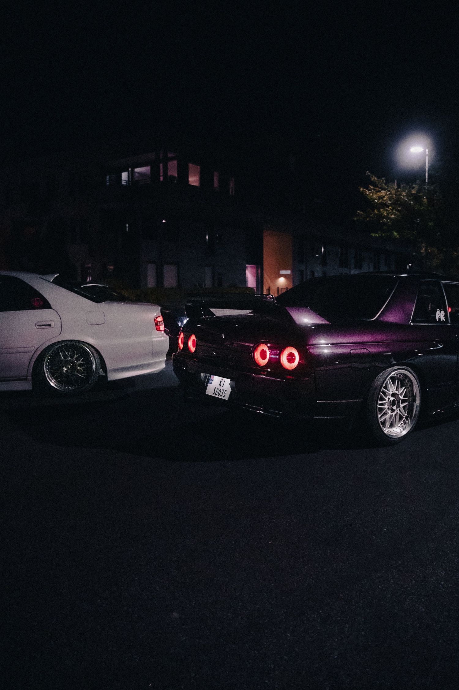 Purple Nissan Skyline R32 and white JDM car parked at night with red tail lights.