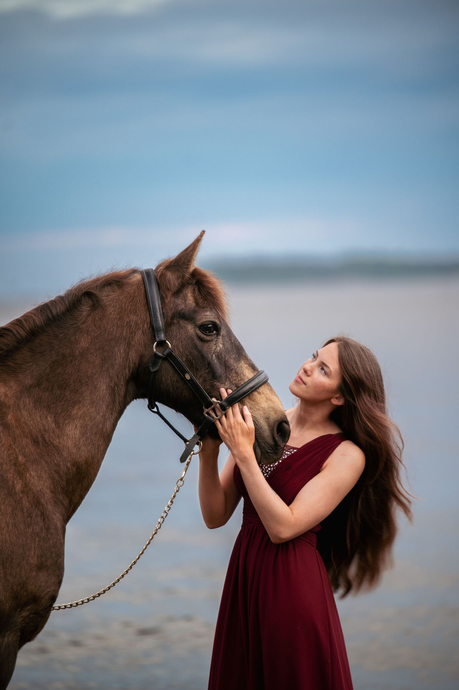 Young woman in burgundy dress tenderly holds the face of a brown horse on a beach.