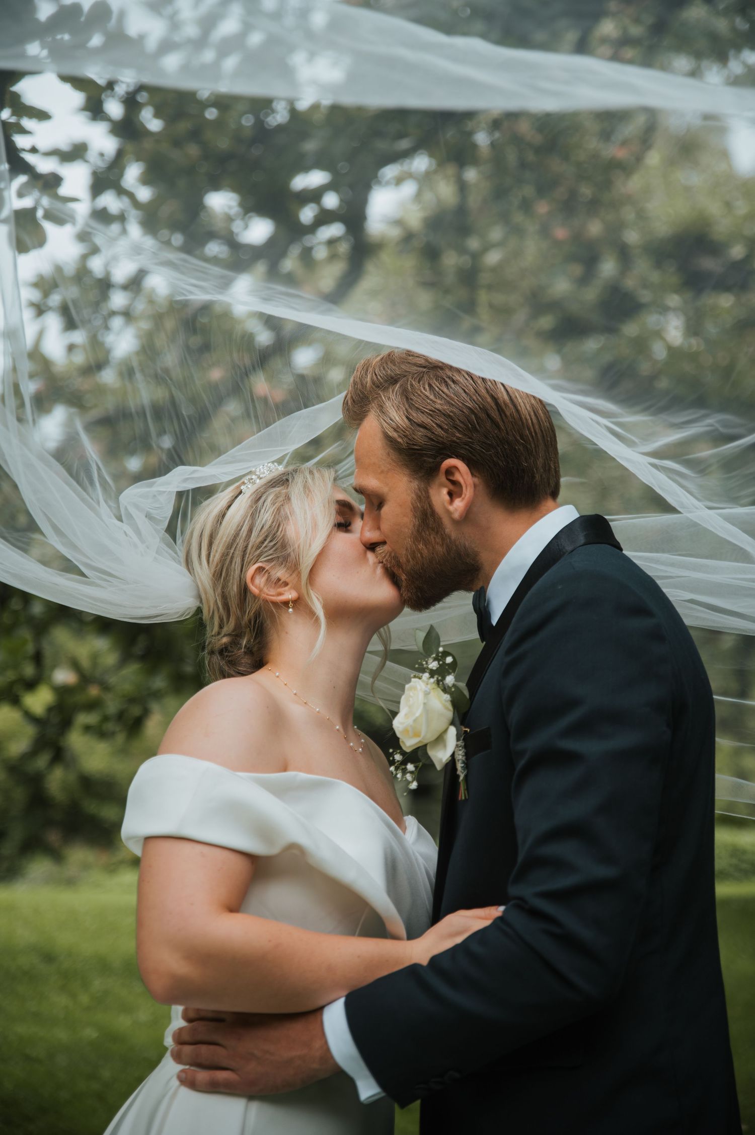 Bride and groom sharing a kiss under a flowing white veil in a garden setting.