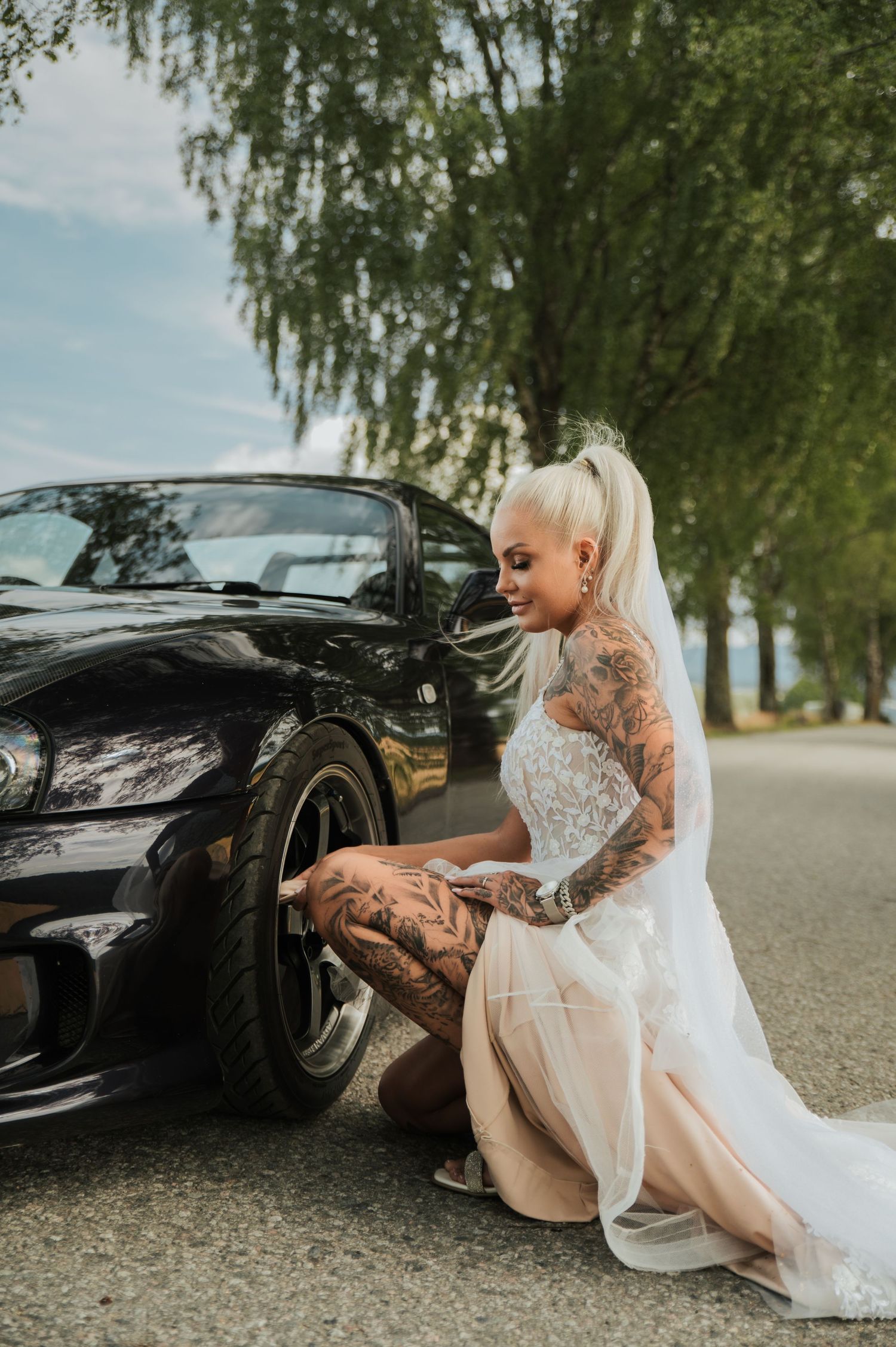 Tattooed blonde bride in white lace wedding dress posing beside a black convertible car.