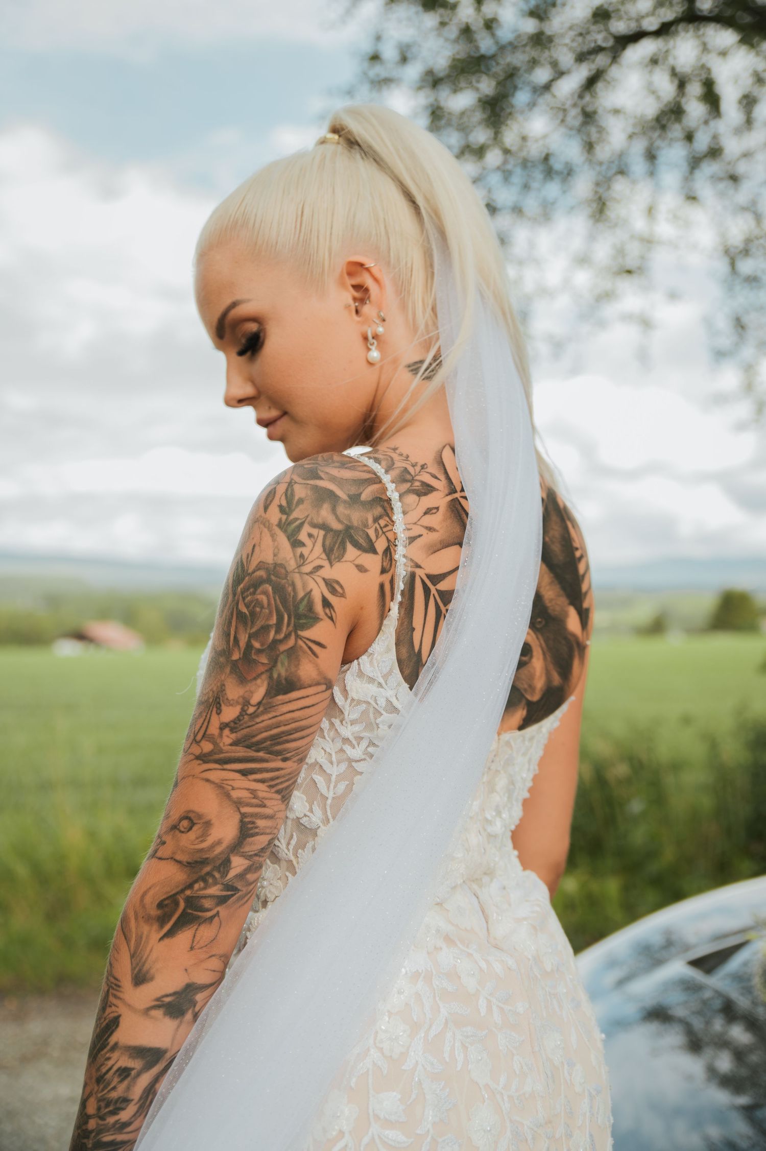Tattooed bride with blonde ponytail in backless white wedding dress with veil outdoors.