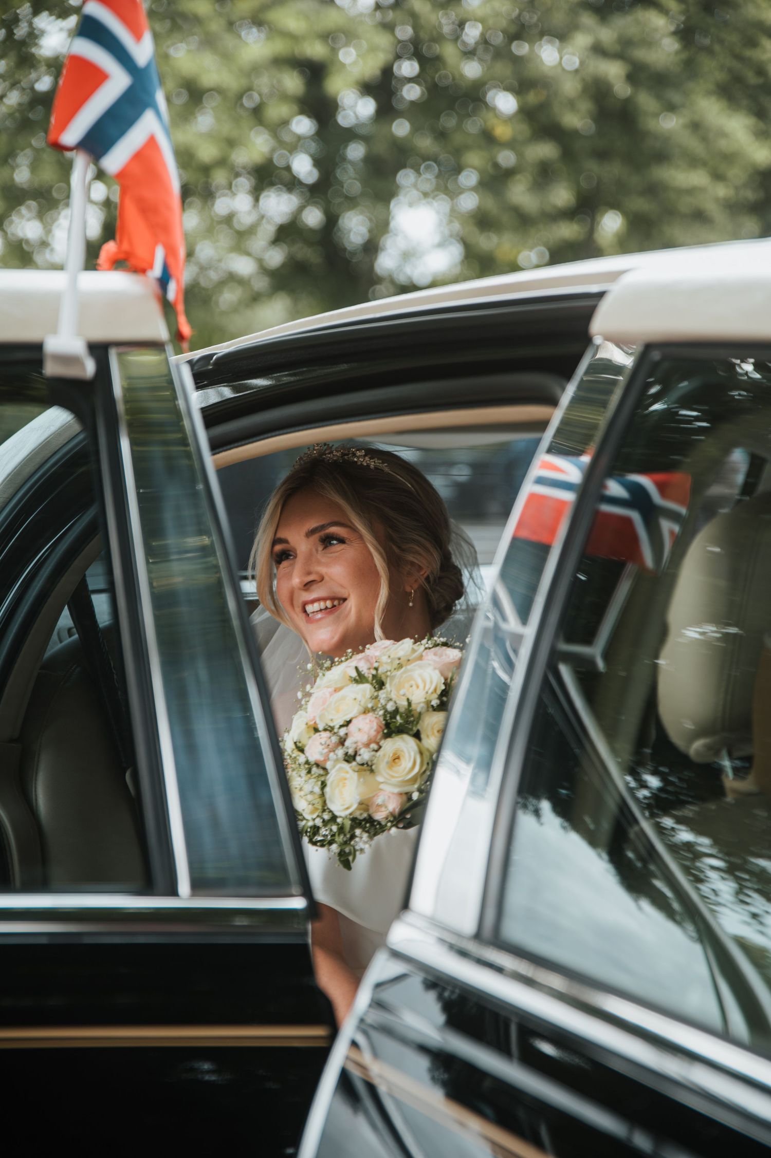 Smiling bride holding a bouquet of white roses stepping out of a decorated car with Norwegian flags.