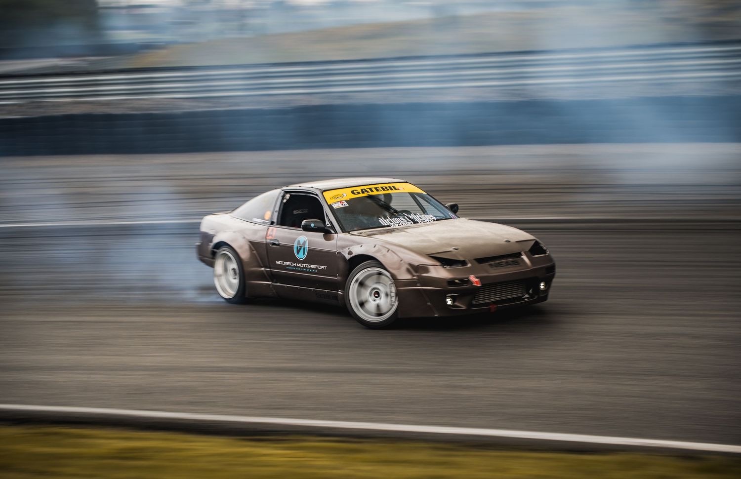Brown Nissan S13 drift car producing smoke while cornering on a race track.