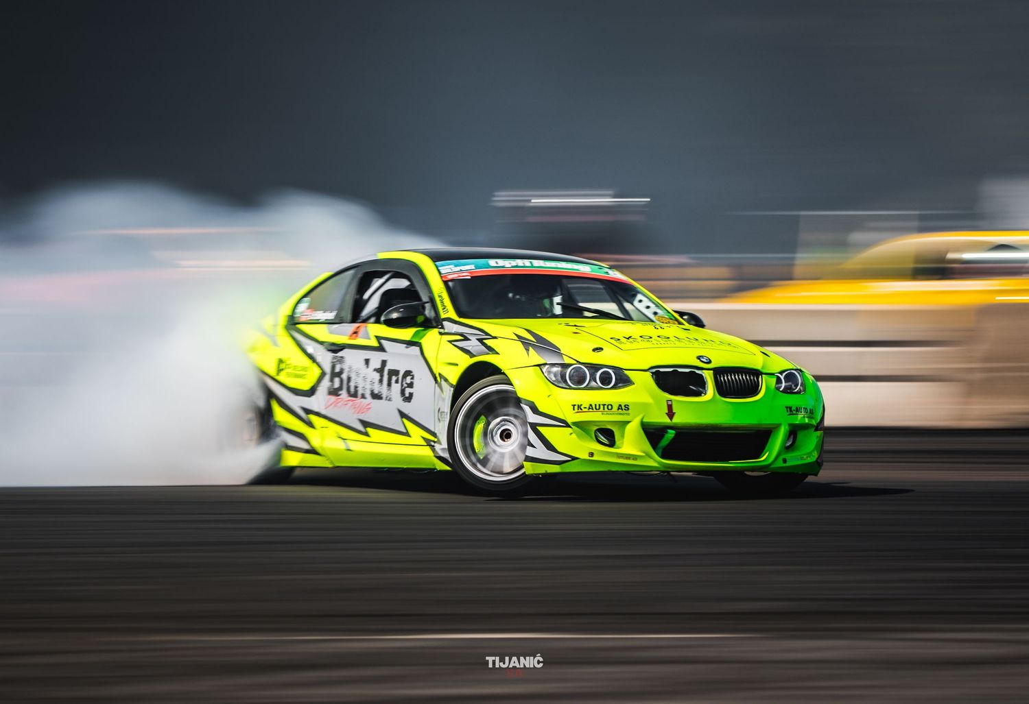 Neon yellow BMW M3 drift car with MaxTire livery producing heavy smoke on competition circuit.