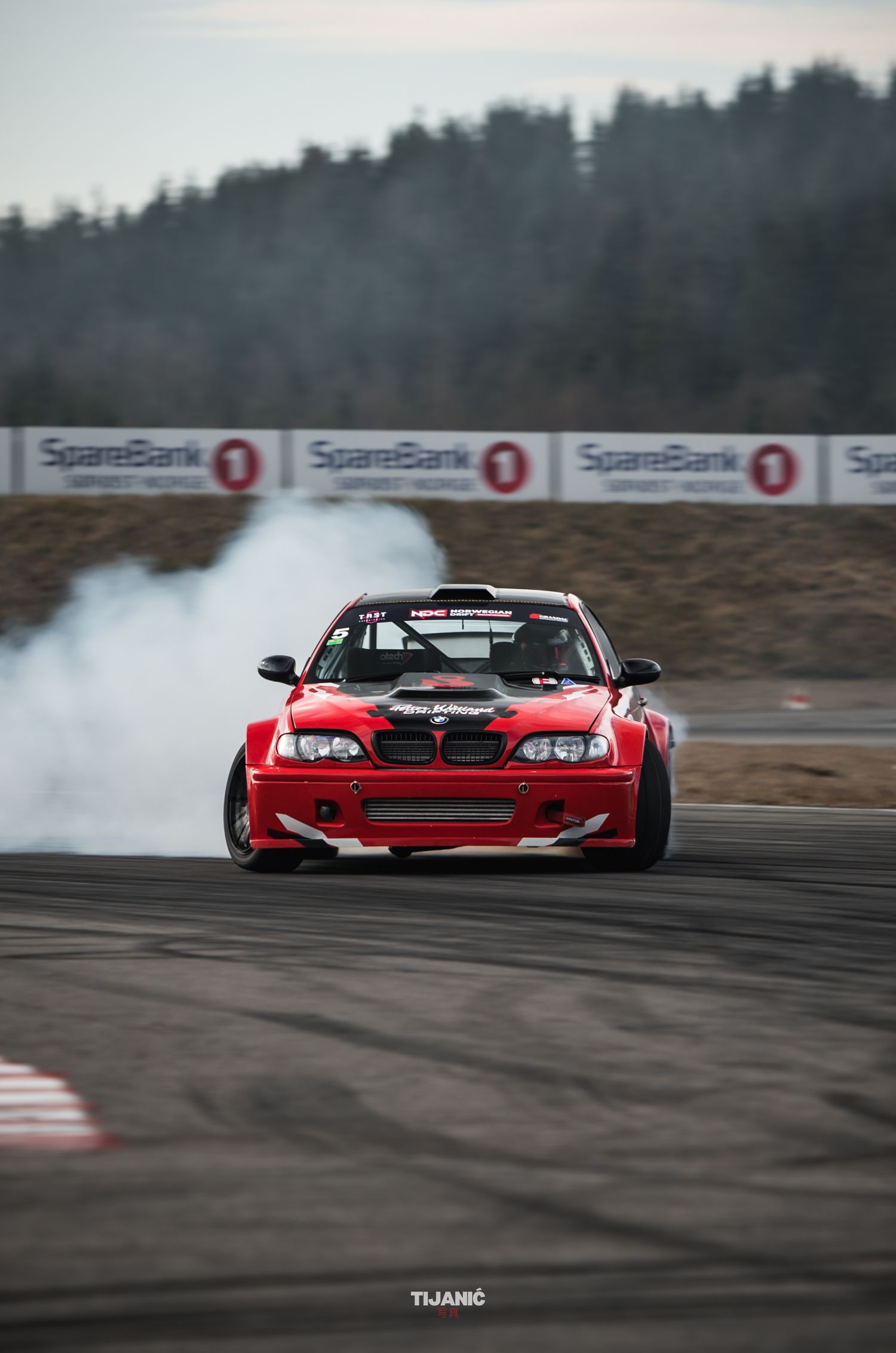 Red BMW E46 drift car generating tire smoke on SpareBank sponsored racing track during competition.