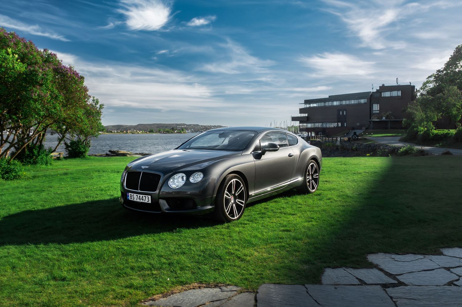 Luxury black Bentley Continental GT parked on green grass beside a modern waterfront property in Norway.