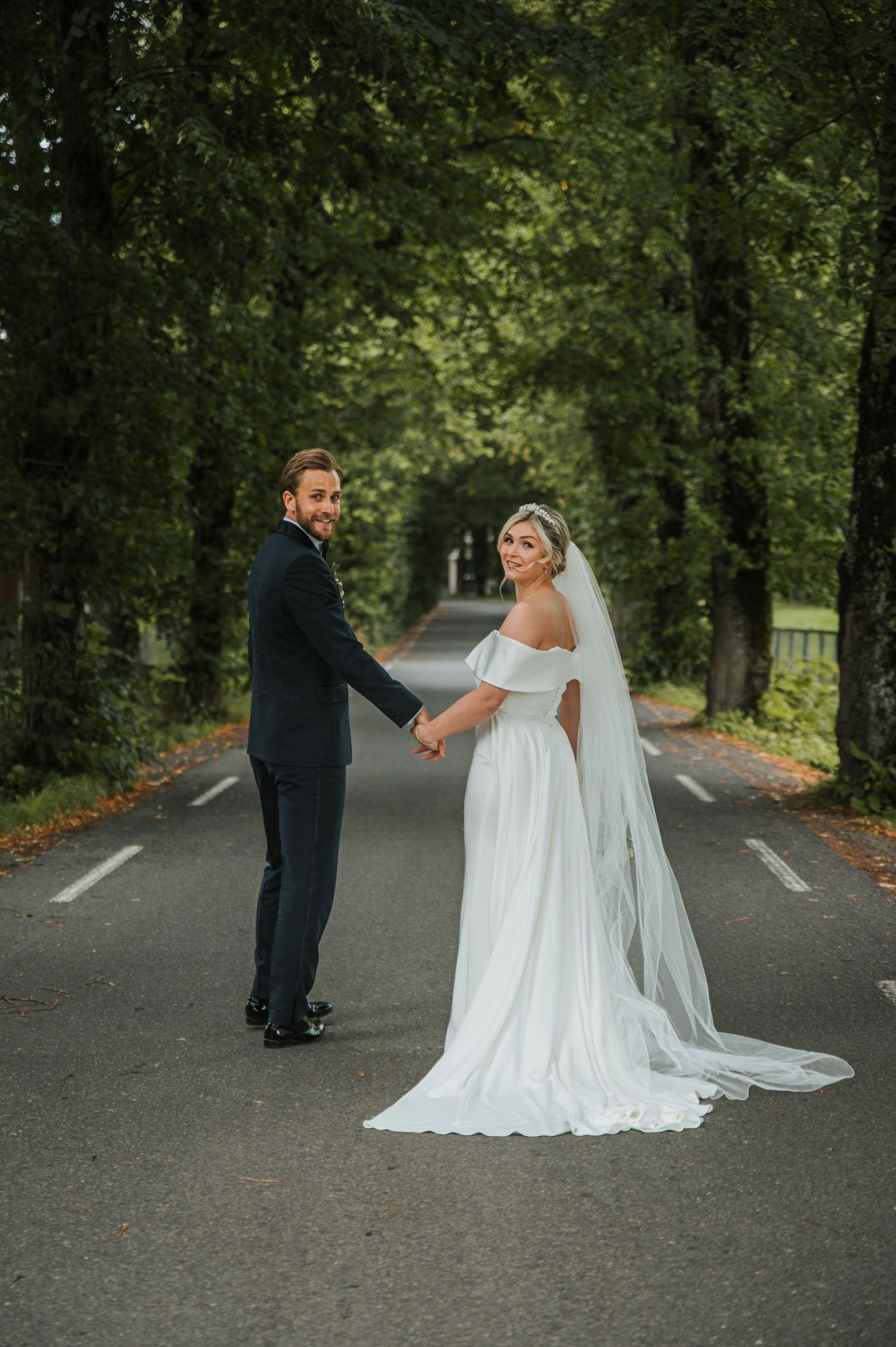 Bride and groom holding hands on tree-lined road, both looking back at camera in elegant wedding attire.