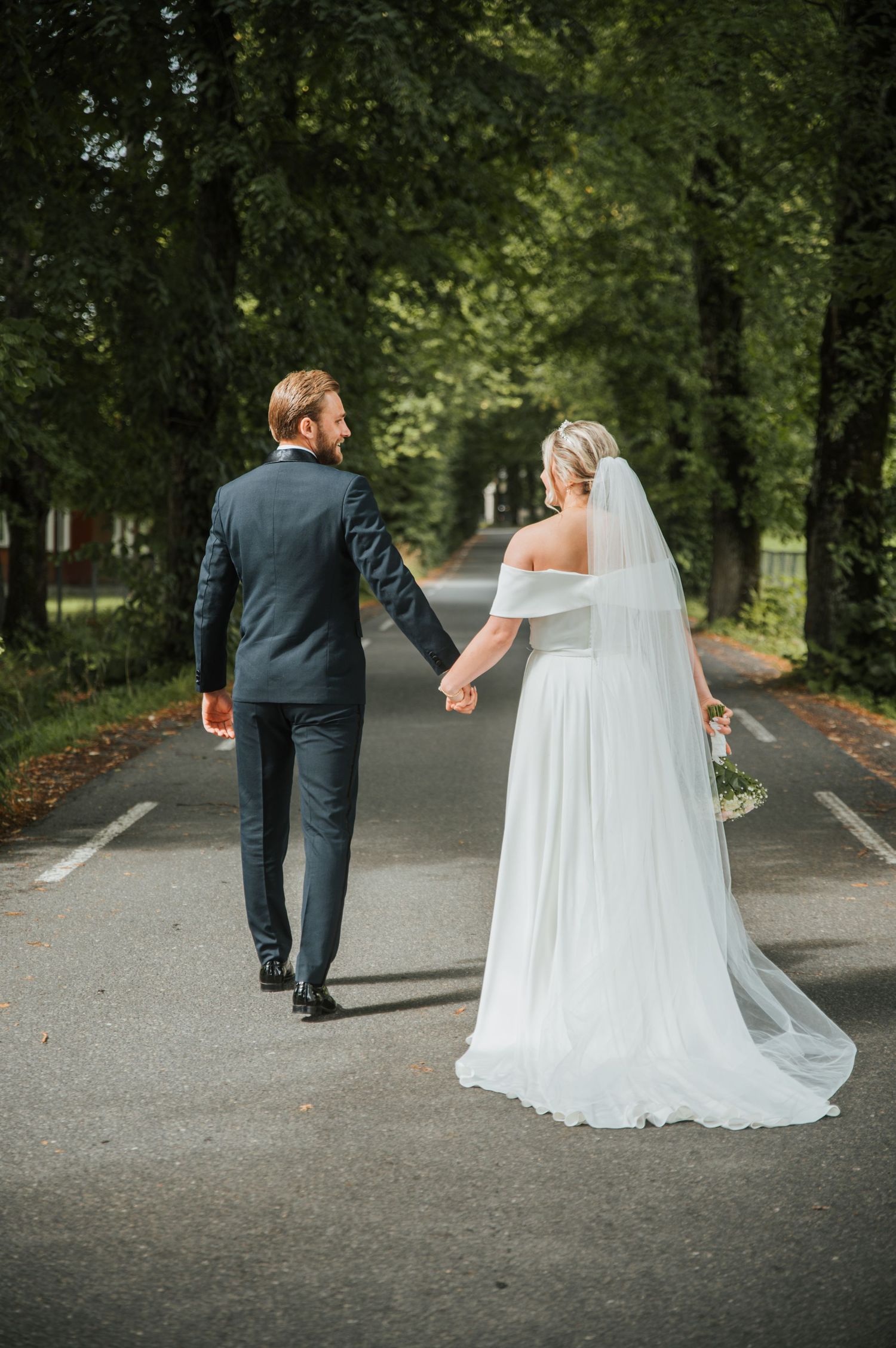 Bride and groom walking hand in hand down a tree-lined road, she in a white gown with long veil.