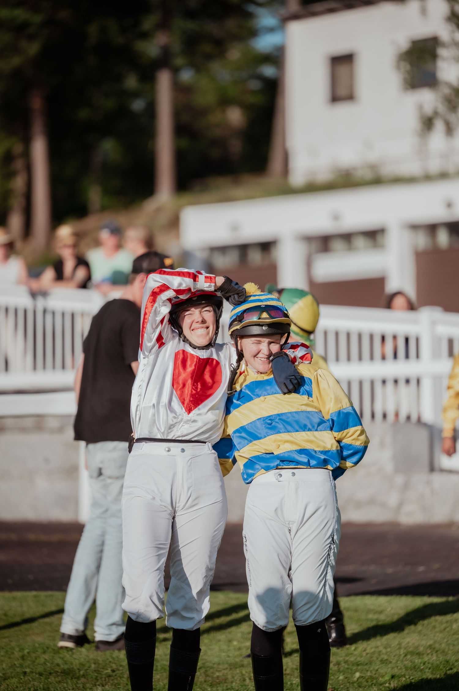 Two smiling jockeys in colorful racing silks standing together at a sunny horse racing track.