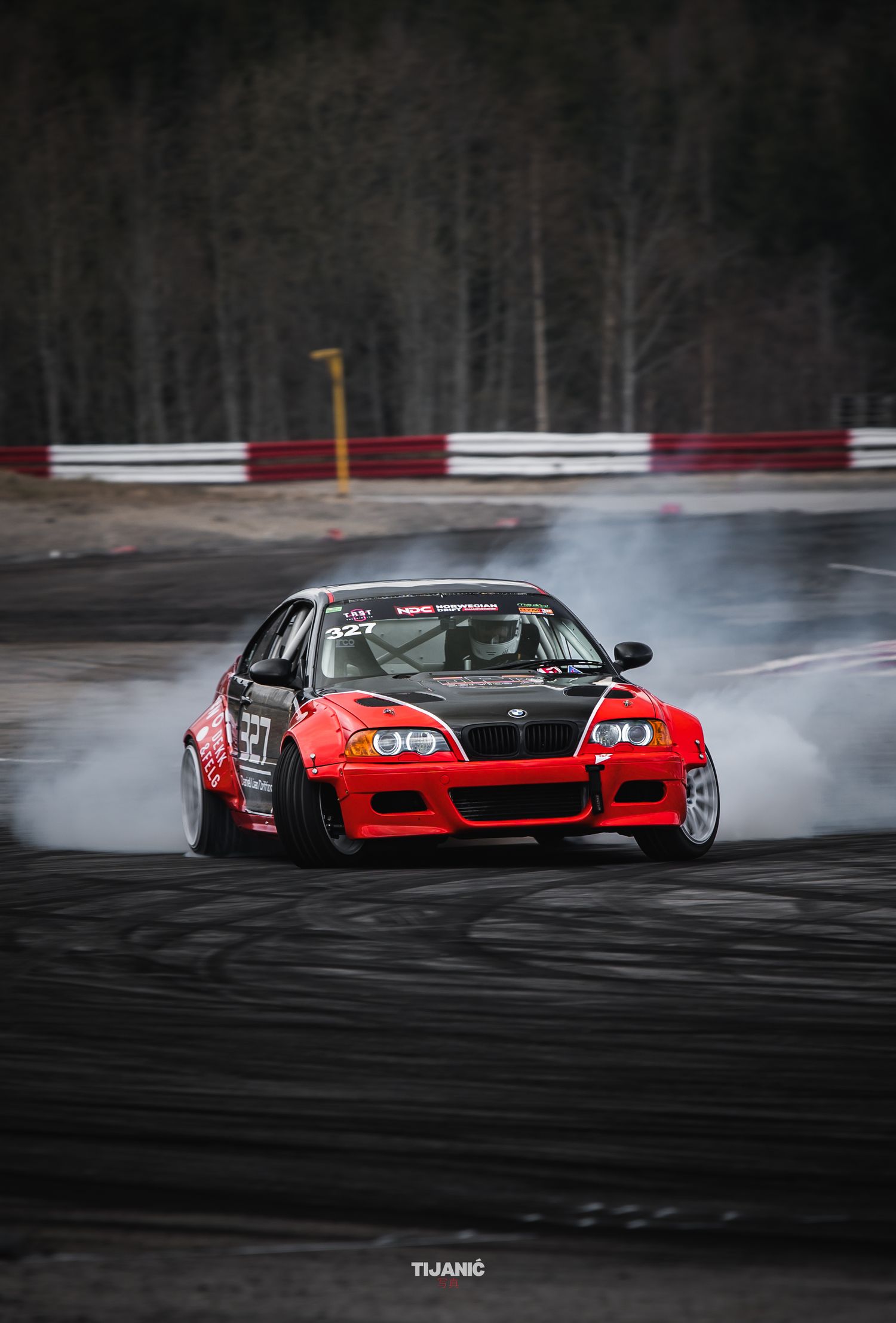 Red BMW drifting on a race track with smoke from tires at a motorsport event.
