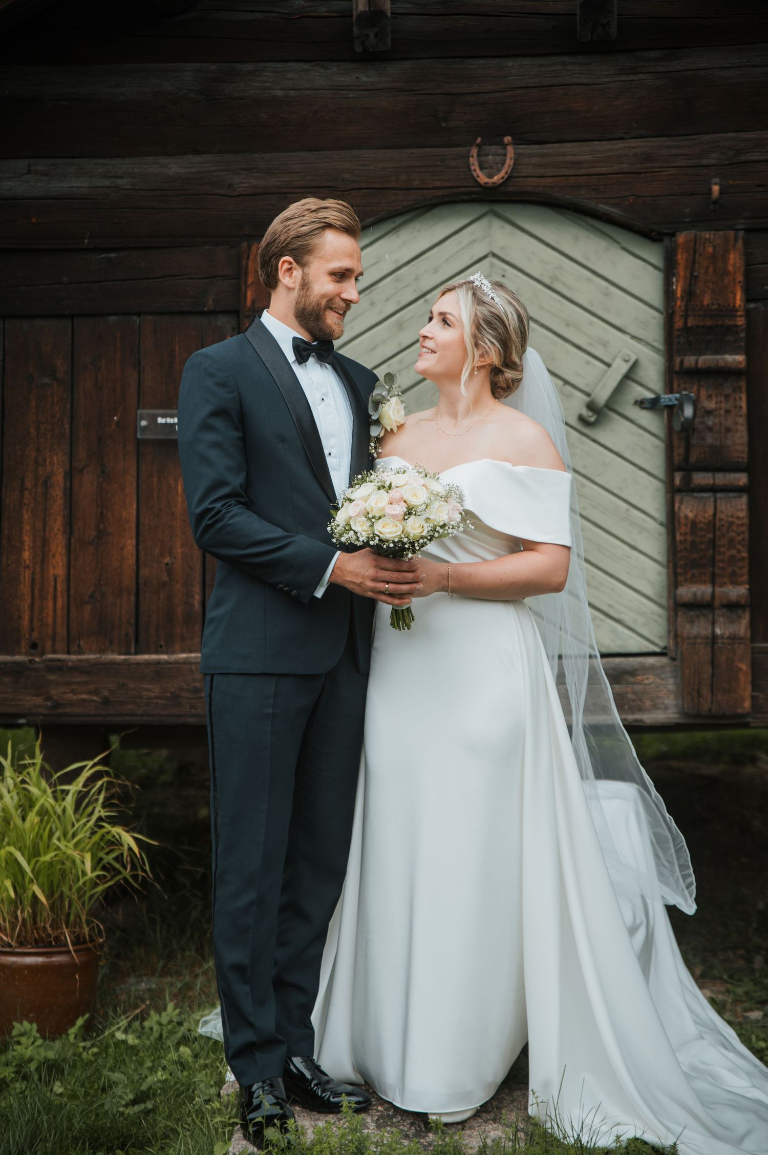 Smiling bride and groom embracing in front of rustic wooden barn, bride holding white rose bouquet.