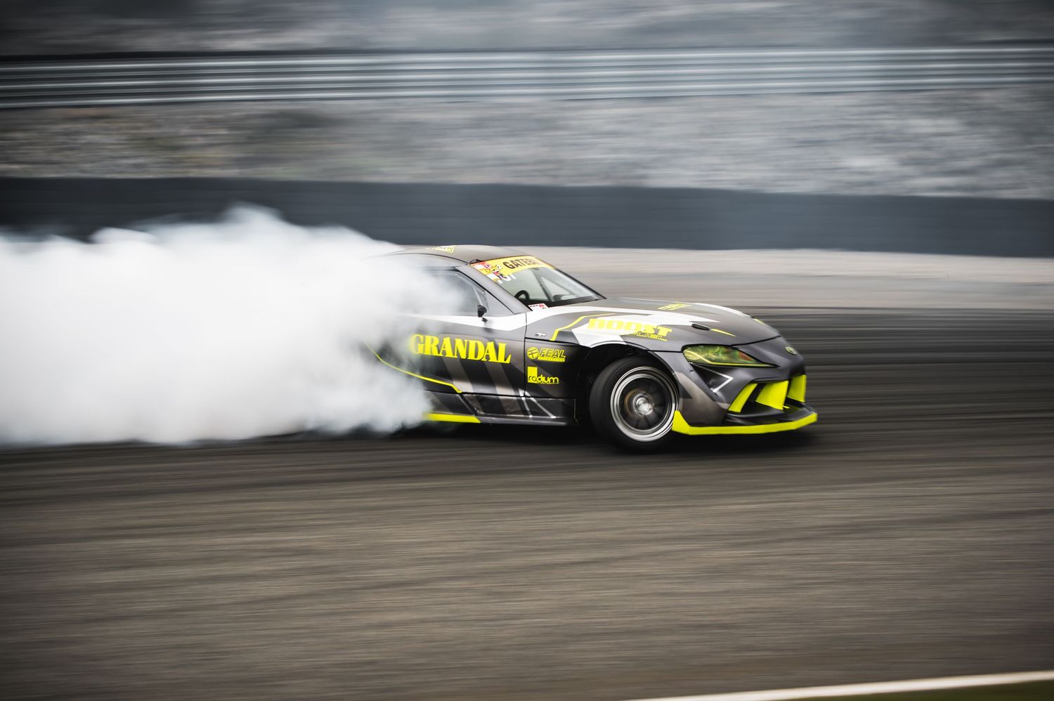 Yellow and black Grandal branded drift car producing massive tire smoke on race track.
