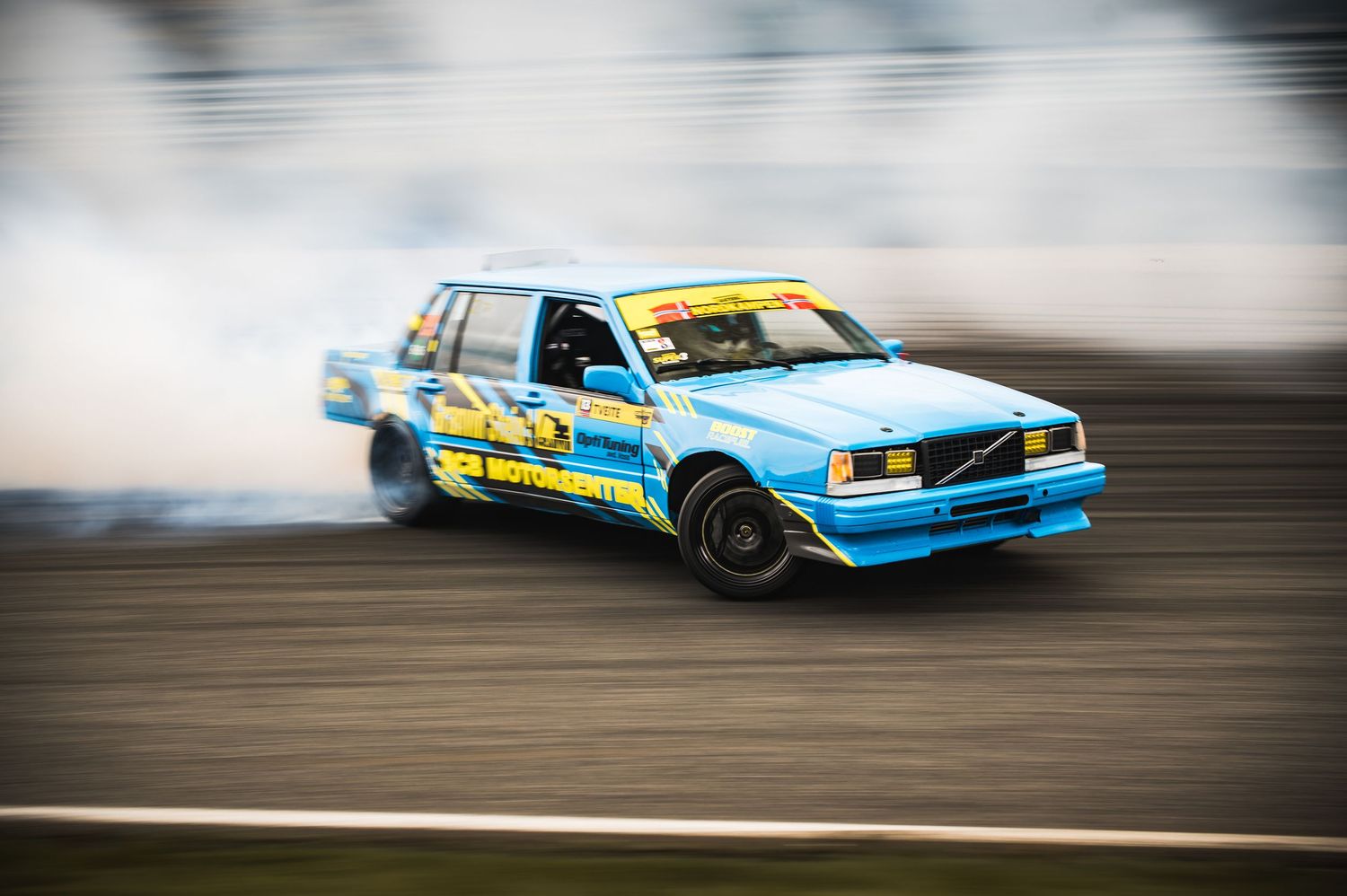Blue Volvo drift car producing tire smoke on a racing track during a drift competition.