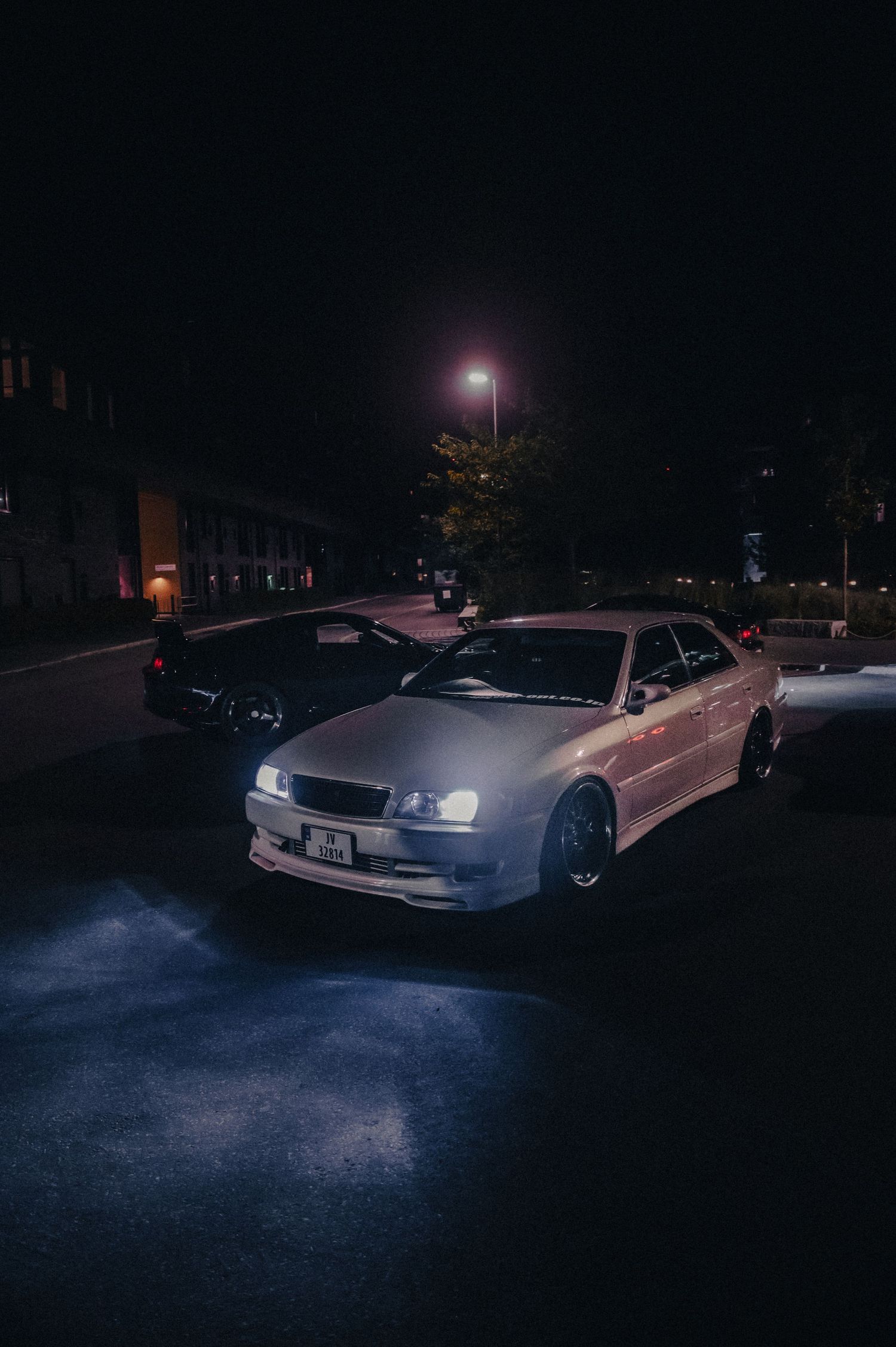 Silver JDM sedan with headlights on parked under a street light at night.