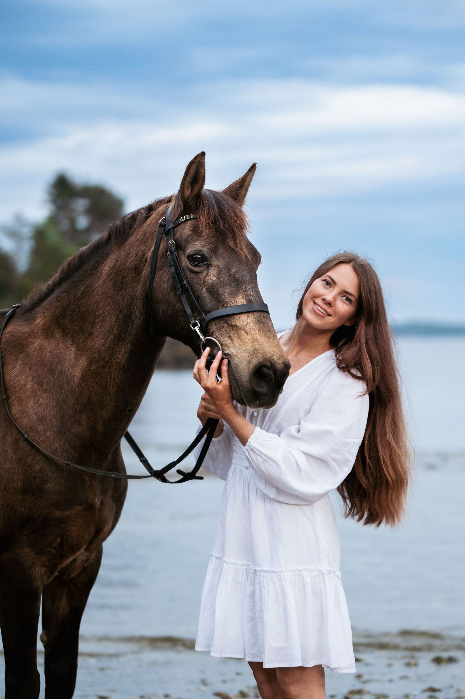 Smiling woman with long hair in white dress poses with a brown horse on a beach.