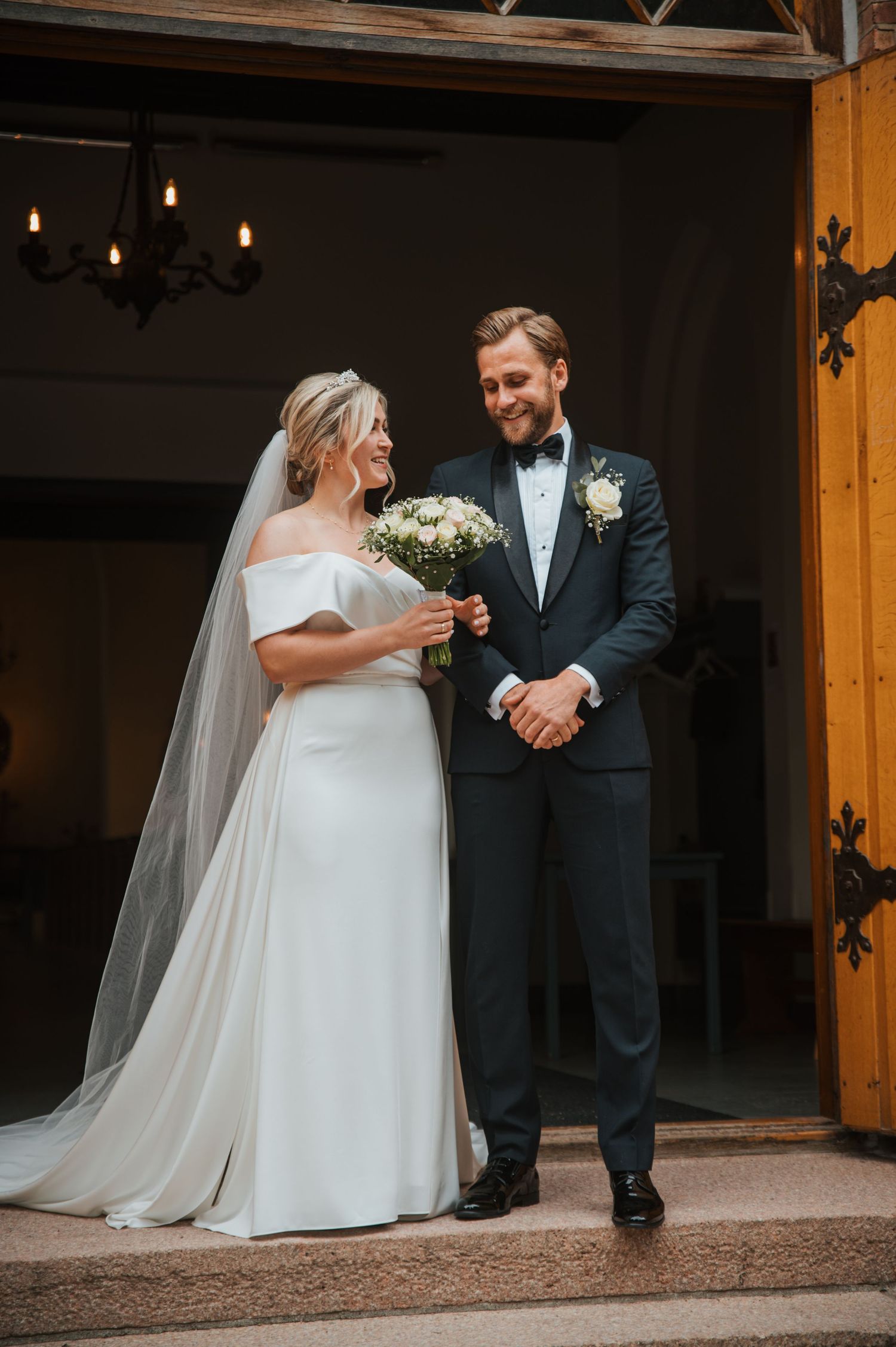 Bride and groom smiling together at church entrance, bride holding white bouquet in elegant off-shoulder gown with veil.