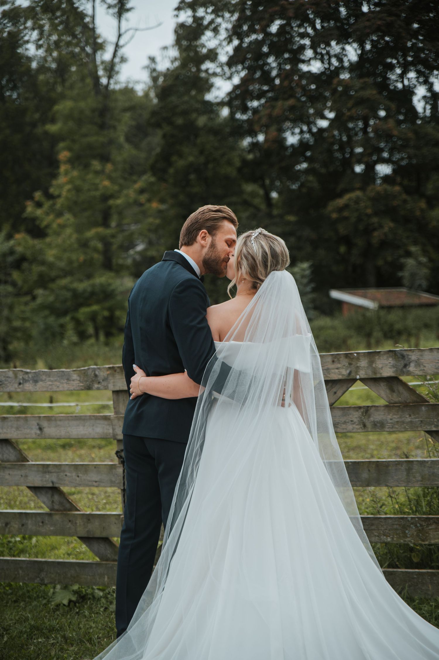 Bride and groom sharing a romantic kiss near a rustic wooden fence surrounded by green trees.