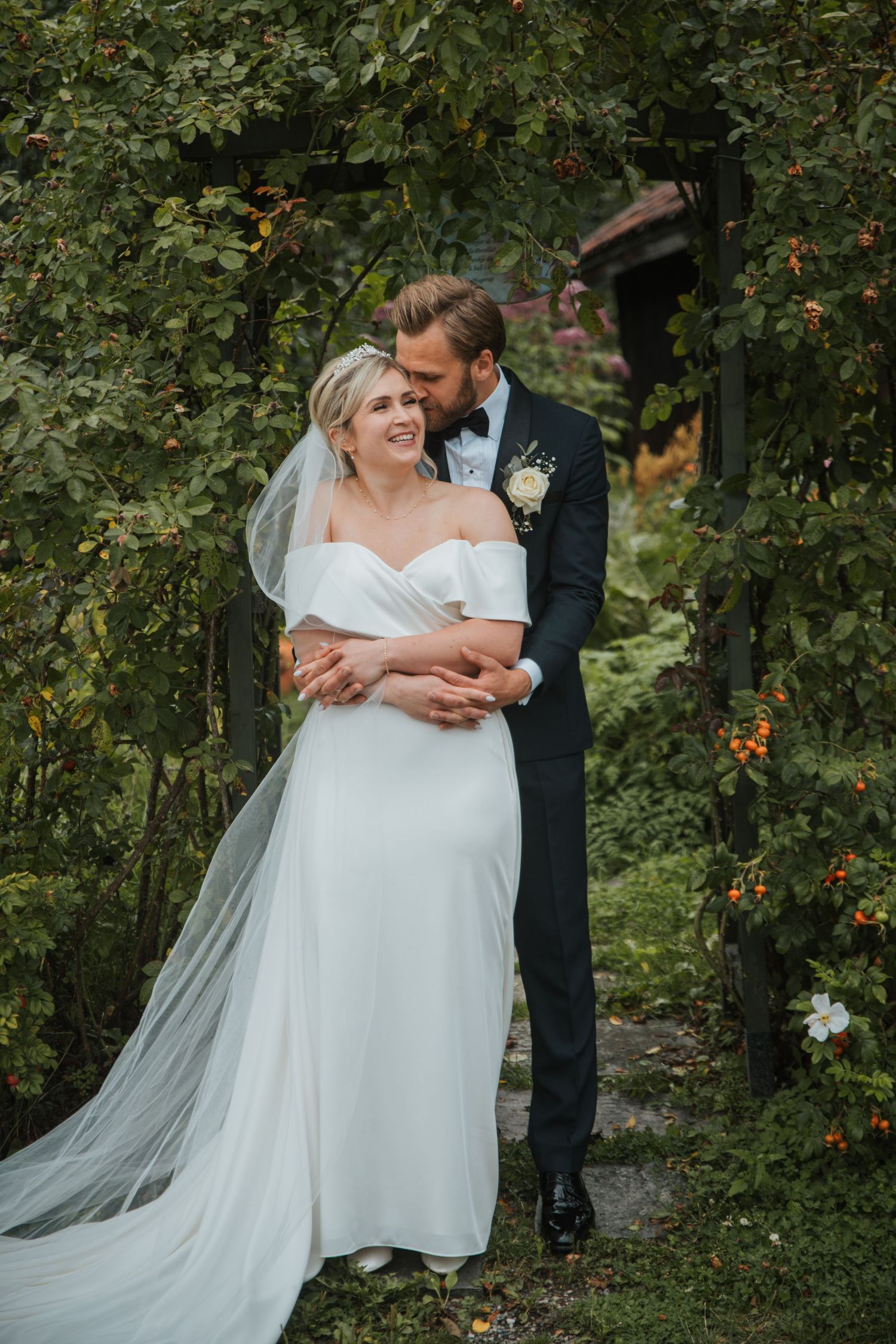 Groom in dark tuxedo tenderly embracing bride in white off-shoulder gown under a lush garden archway.