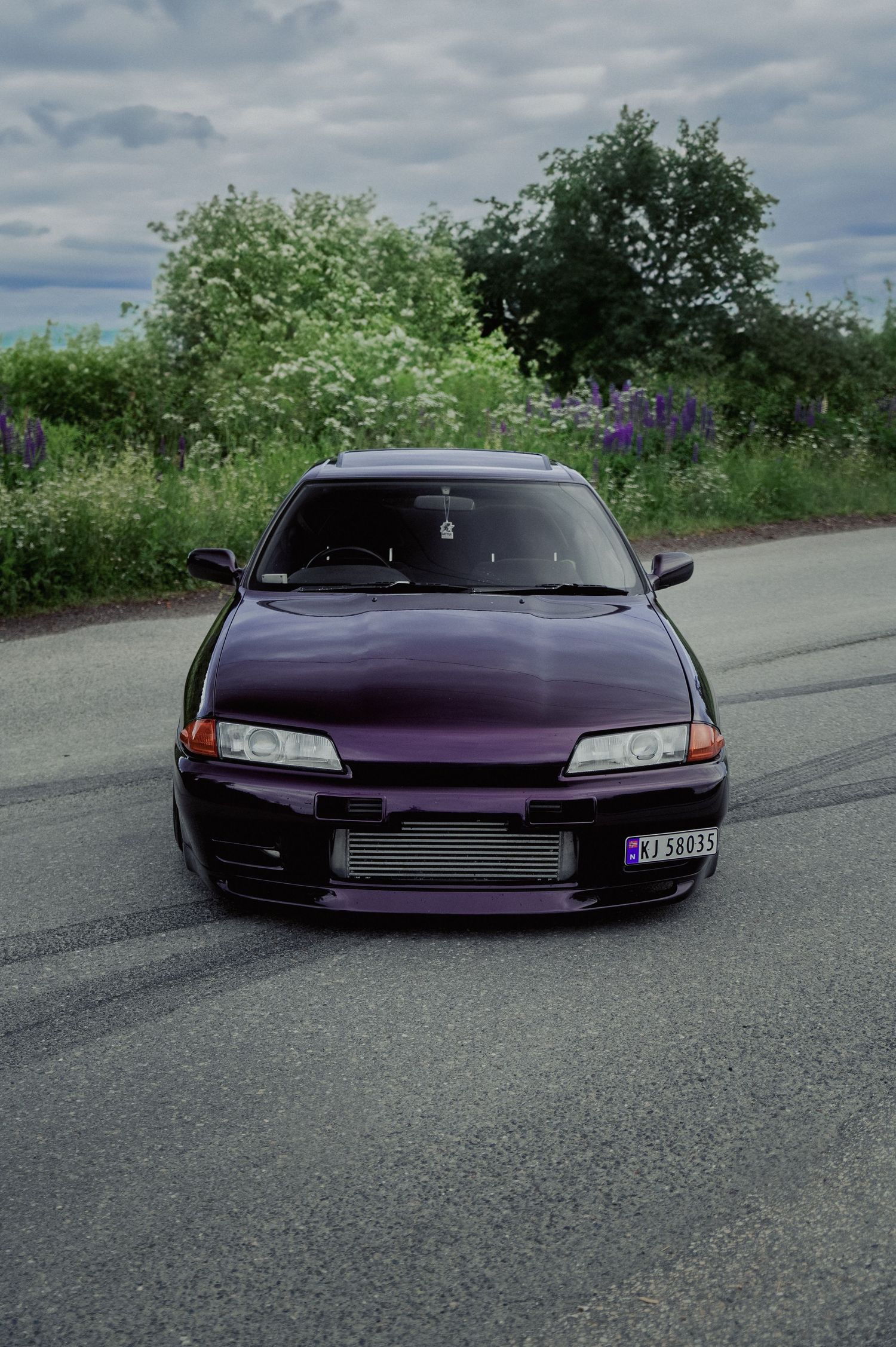 Purple modified Nissan Skyline R32 GT-R front view parked on a road with green trees in background.