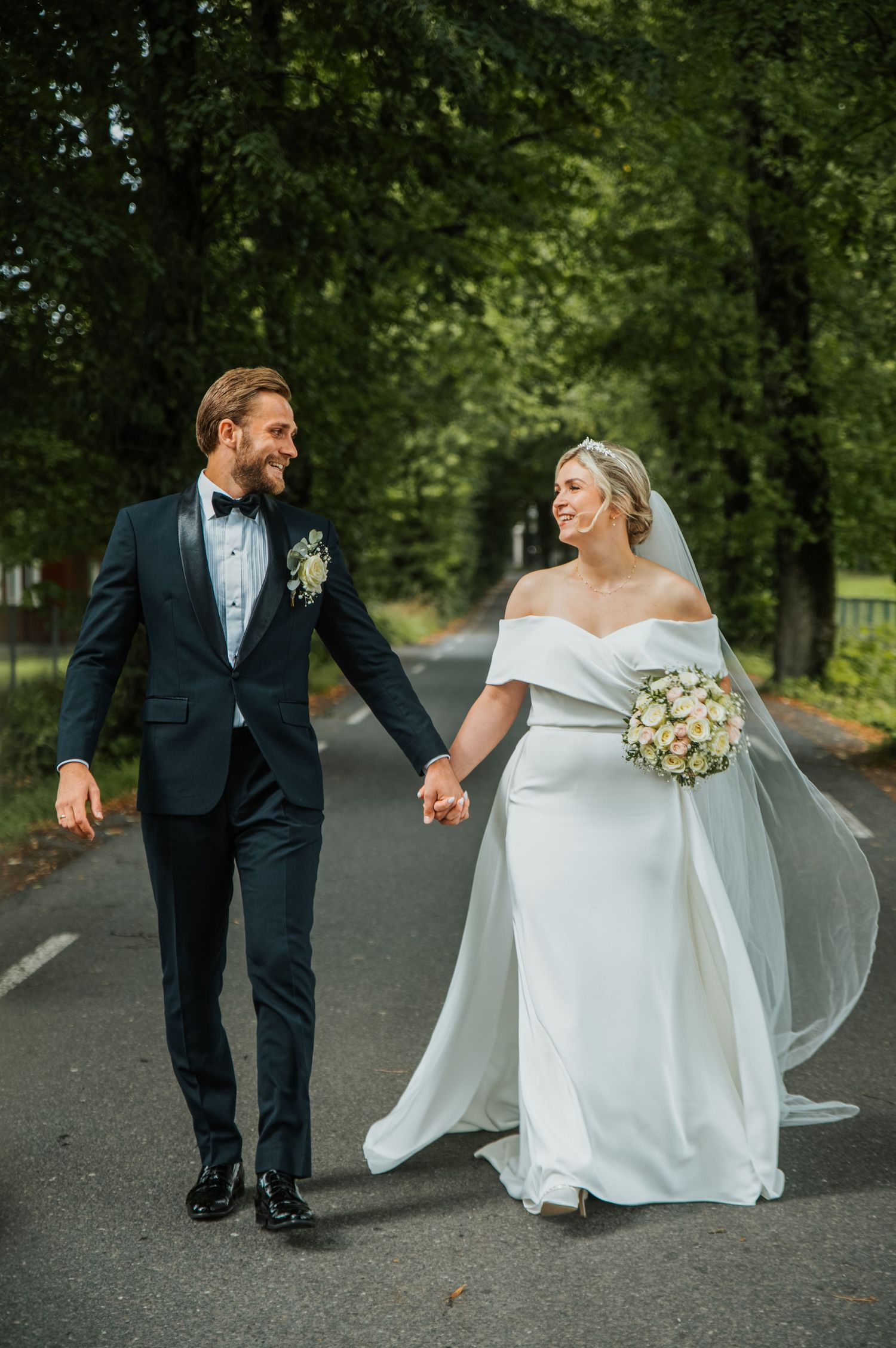 Happy bride and groom holding hands walking on tree-lined road, bride in off-shoulder gown with flowing veil.