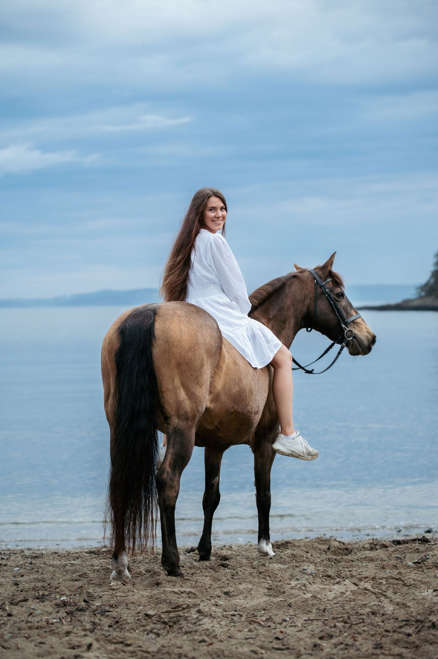Smiling woman in white dress rides a brown horse bareback along a scenic beach.