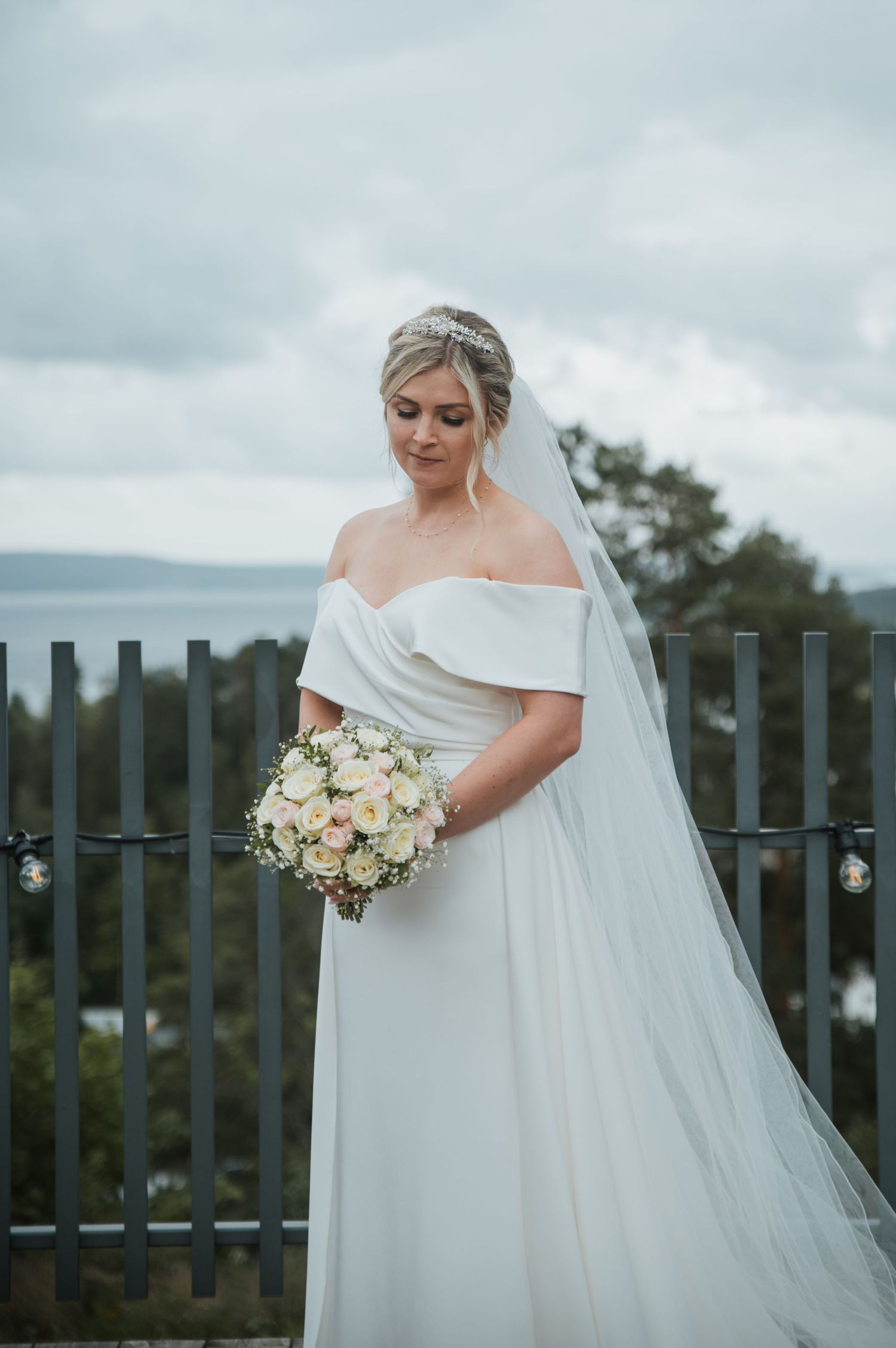 Bride in off-shoulder white wedding gown holding pastel rose bouquet on an outdoor terrace with scenic view.