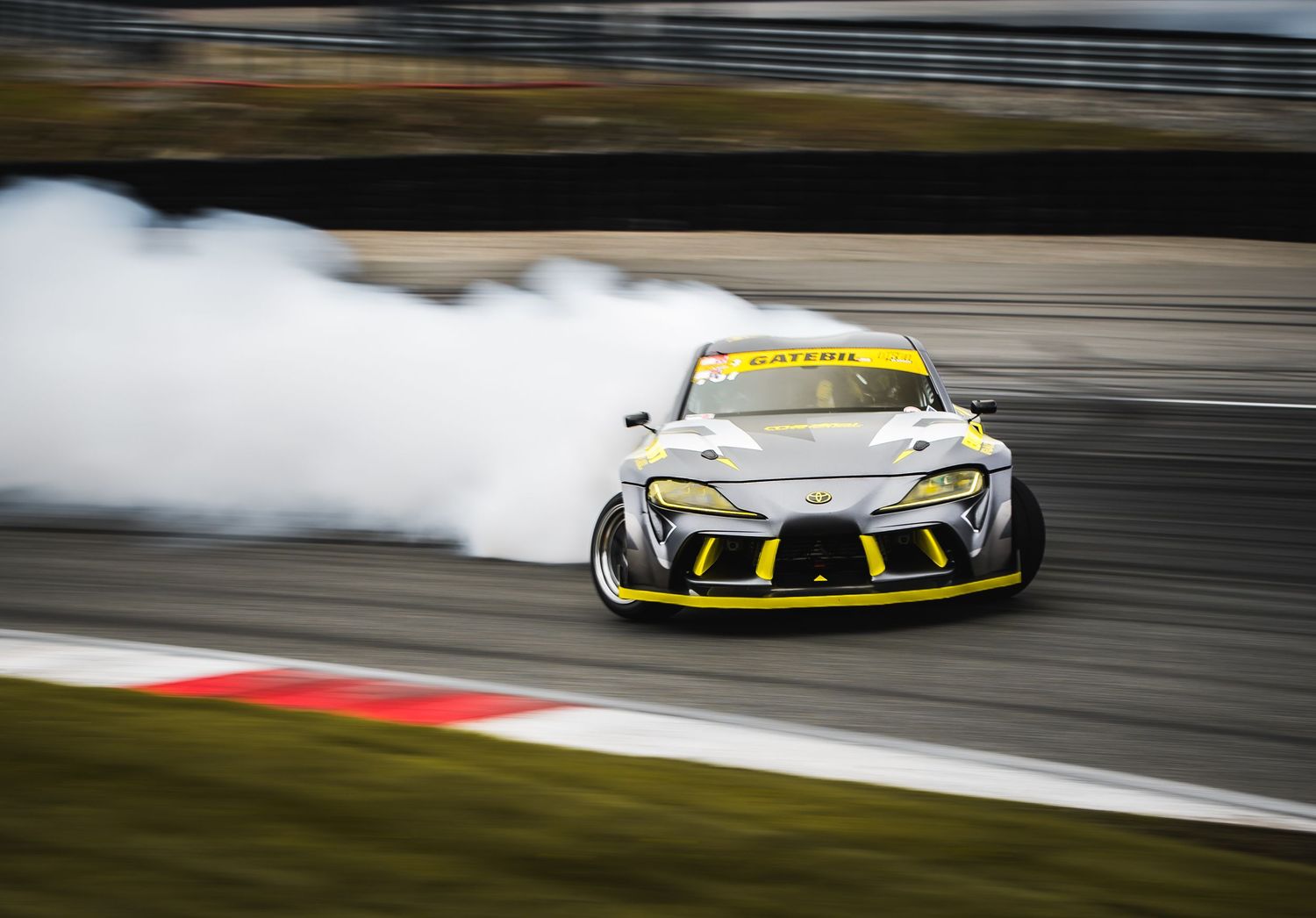 Yellow and grey Toyota Supra drift car producing heavy tire smoke on racing circuit.