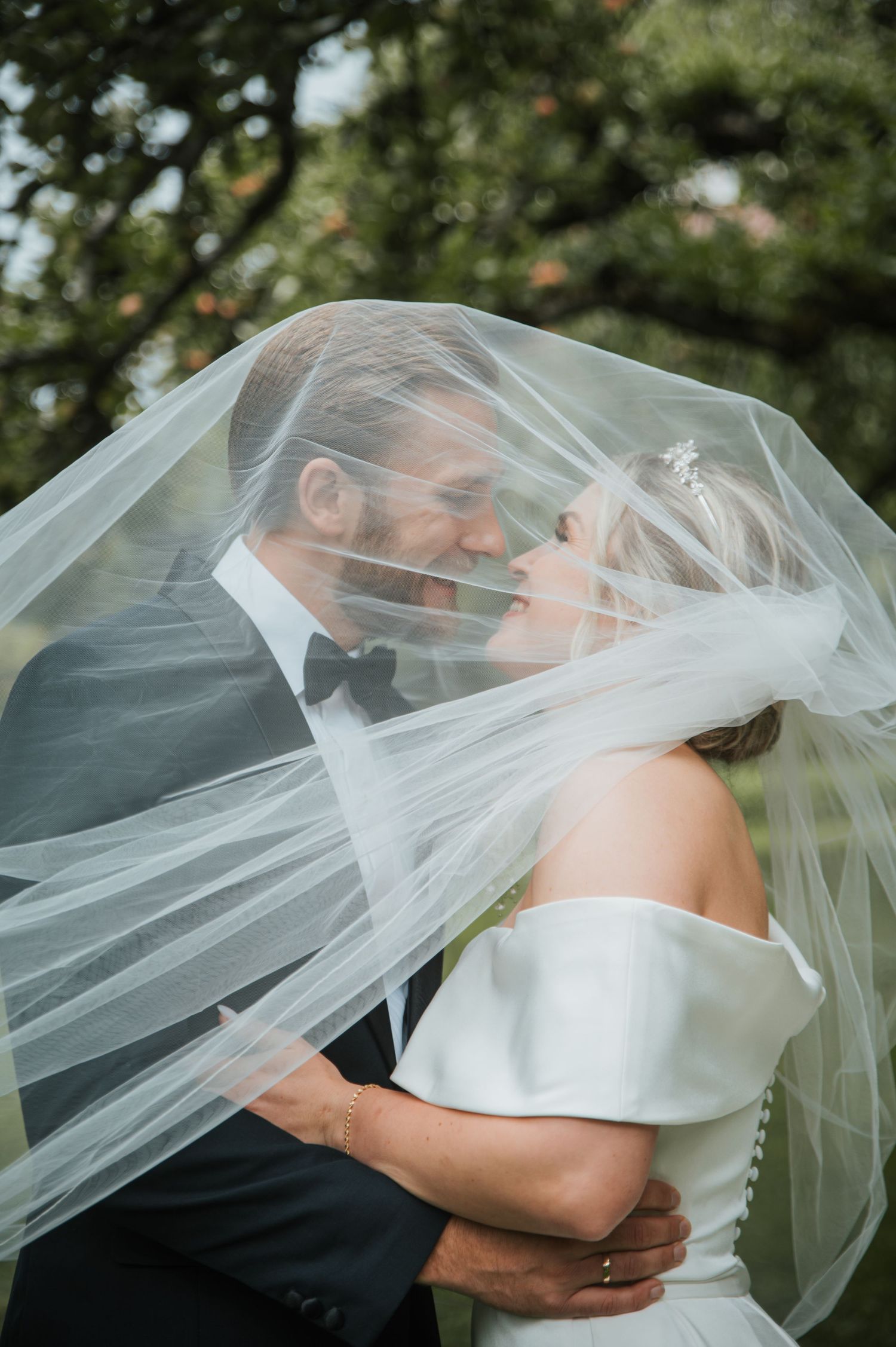 Romantic bride and groom sharing intimate moment under flowing wedding veil outdoors surrounded by greenery.