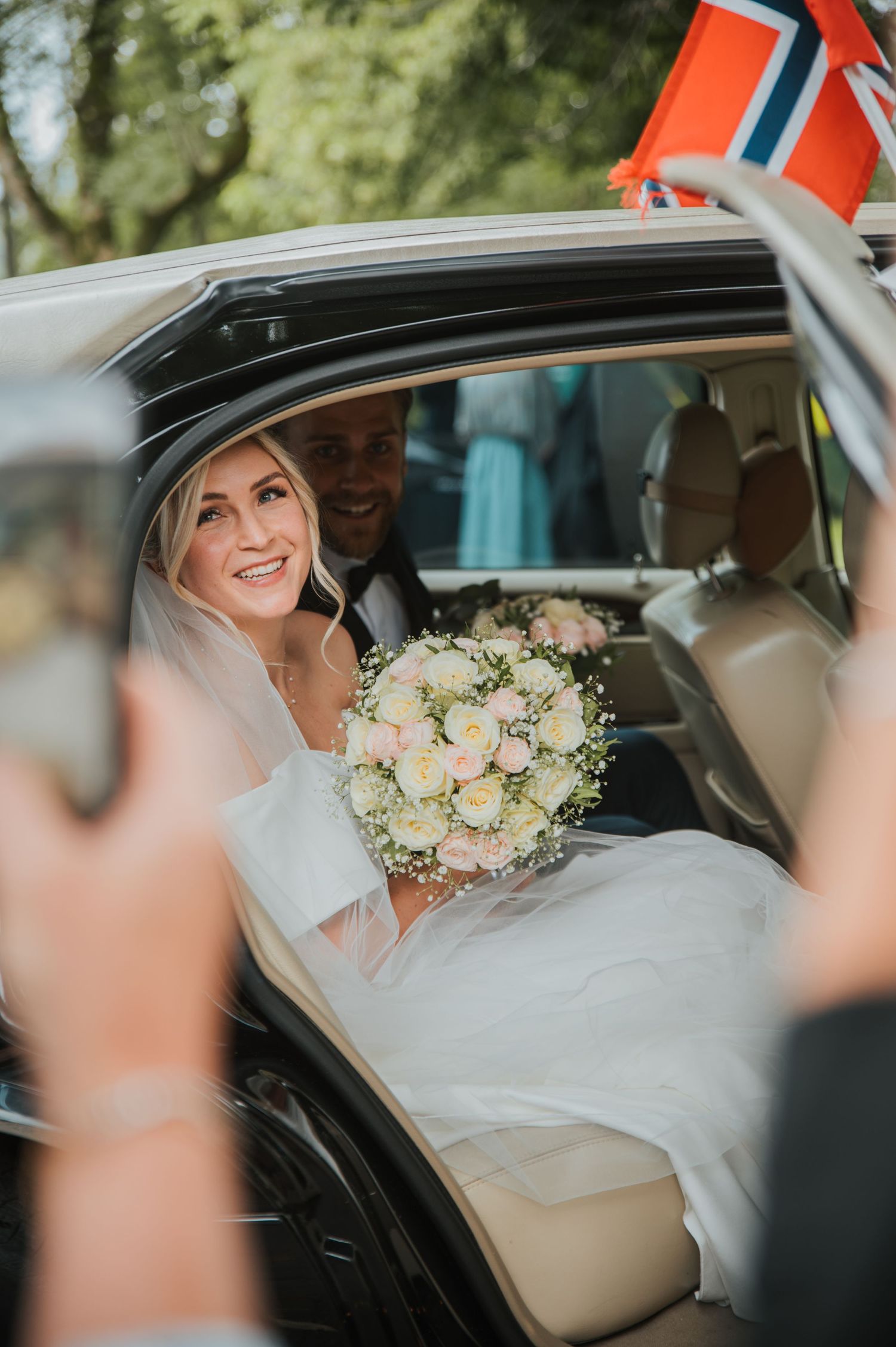 Smiling bride holding bouquet of roses sitting in a wedding car decorated with Norwegian flag.